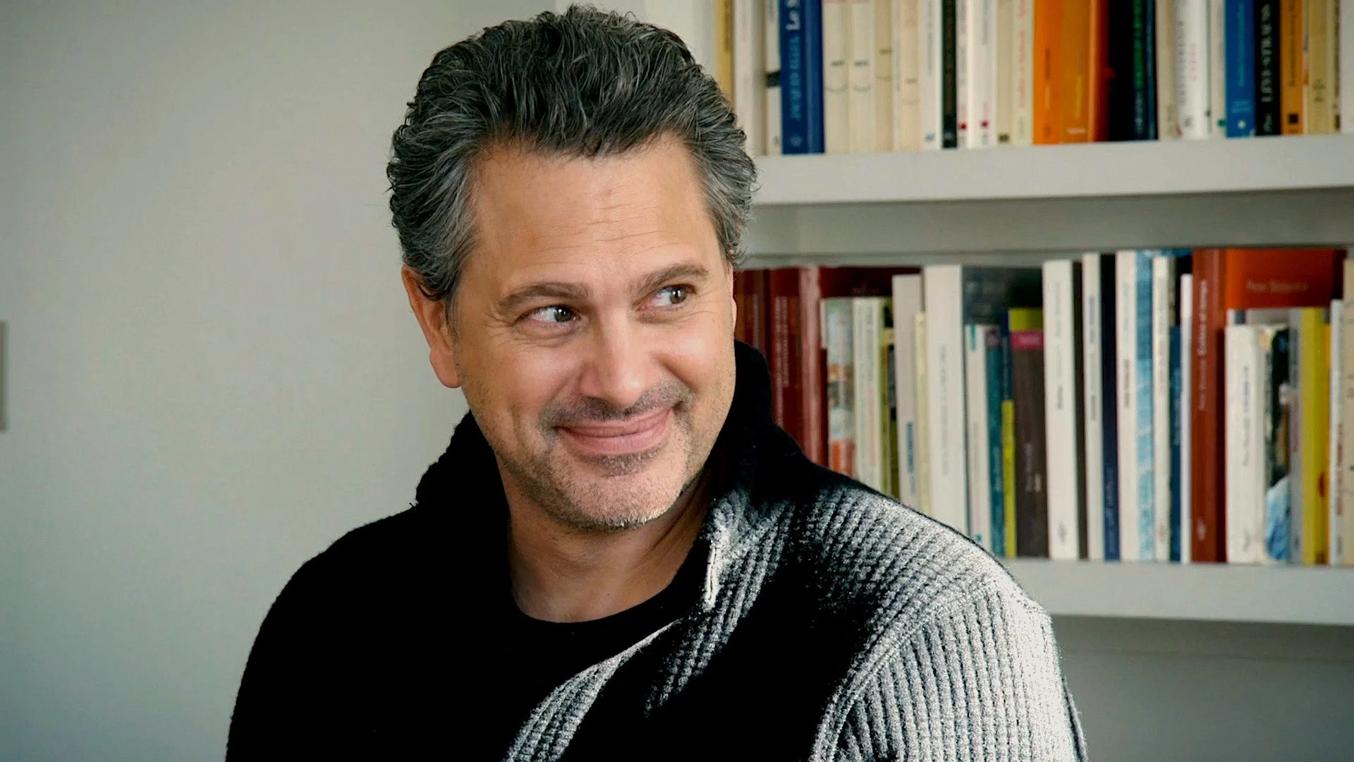 A man with dark, wavy hair and stubble, smiling and looking to the side, sitting in front of a bookshelf filled with books.