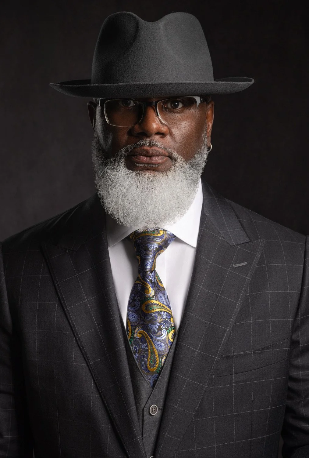 A distinguished African American man with a white beard, wearing a dark checkered suit, white shirt, patterned tie, black hat, and glasses, against a dark background.