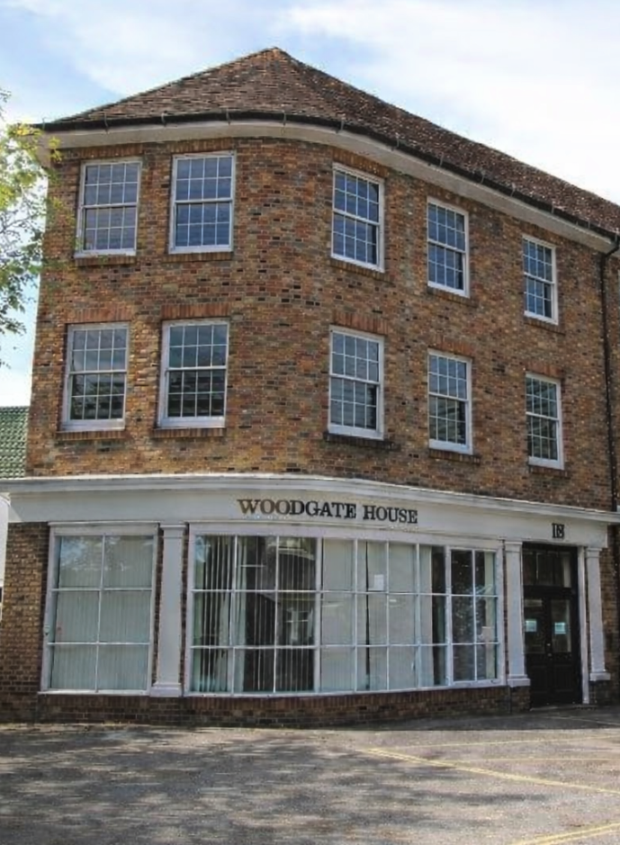 This image shows a three-story brick building named 'Woodgate House' with large glass windows on the ground floor and smaller windows on the upper floors, under a partly cloudy sky.