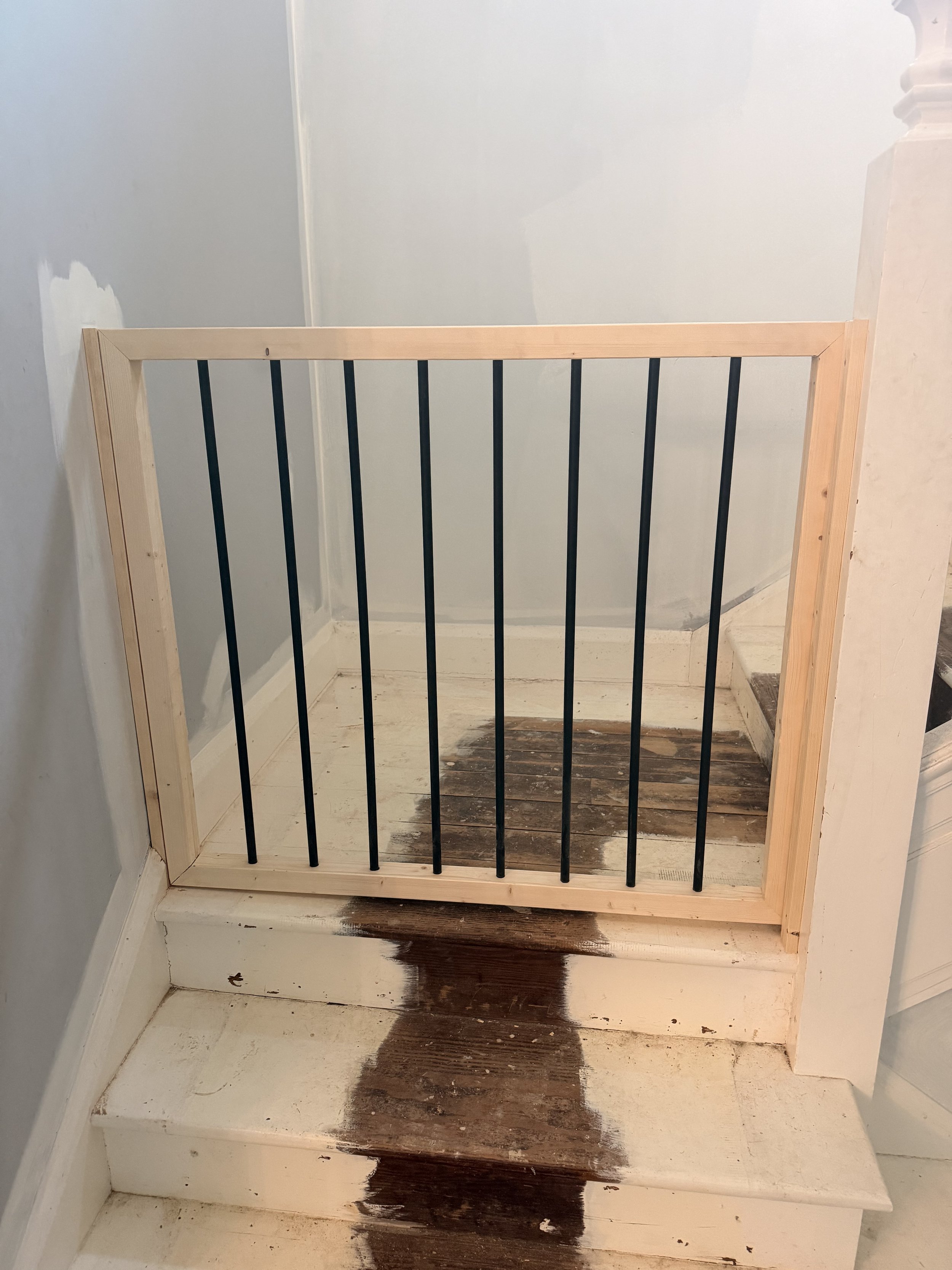 Wood-framed safety gate installed at the top of a stairway, with black metal bars and a white painted staircase below. The staircase has some stained areas on the steps.