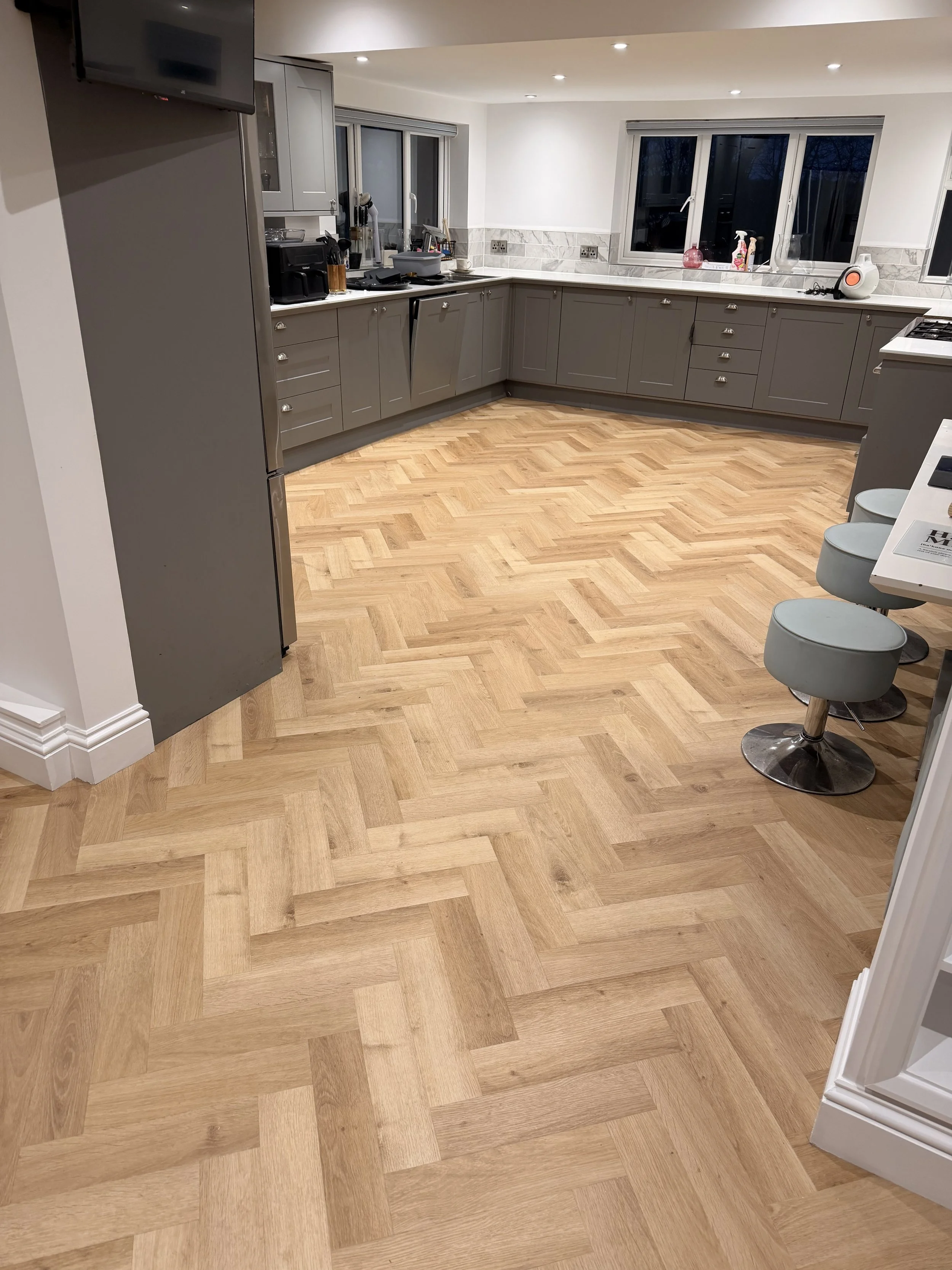 Kitchens with wooden parquet floors, gray cabinets, a marble countertop, and windows. There are some kitchen appliances and a small breakfast bar with stools.