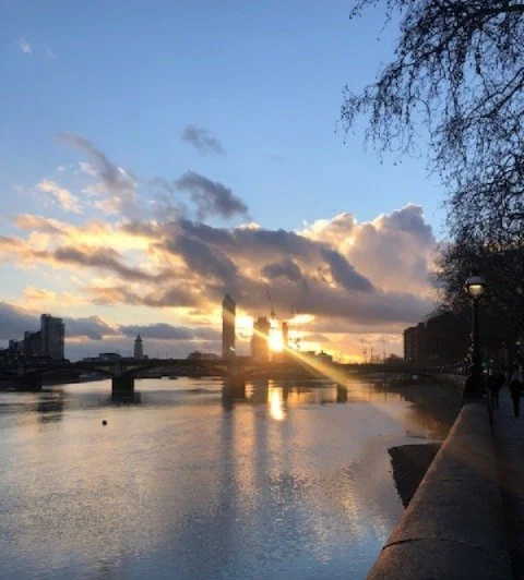 Sunset over a river in an urban setting with clouds in the sky, buildings, and a bridge in the background.