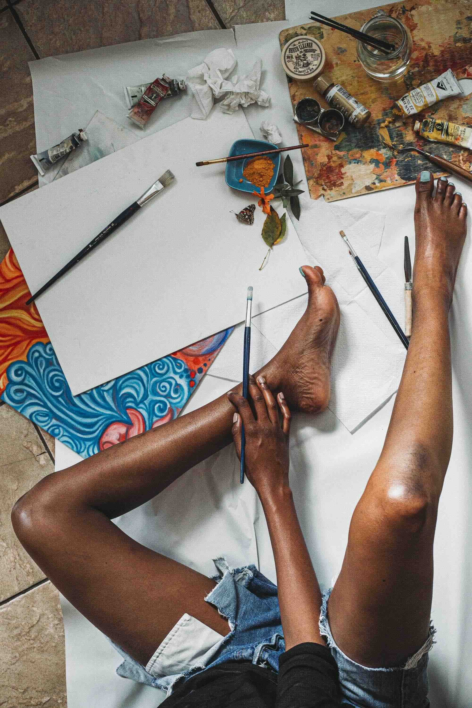 Une personne trépointe à l'envers, en shorts en denim, en train de peindre ou de dessiner avec des pinceaux, entourée de divers tubes de peinture, de feuilles blanches, d'un verre d'eau, et d'autres fournitures artistiques sur une table.