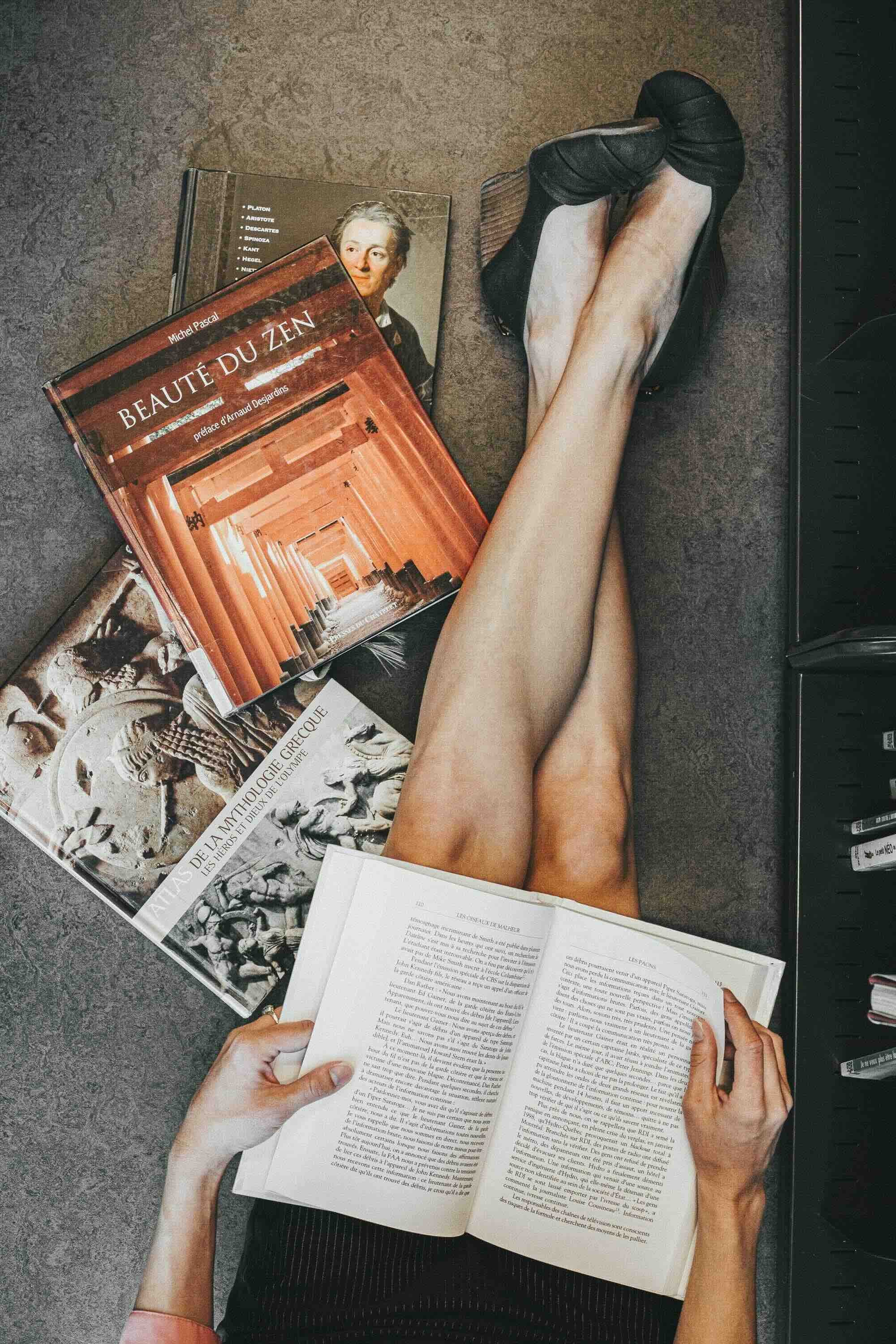 Jambes de femme portant des chaussures à talons hauts, lisant un livre, entourée de plusieurs autres livres sur la philosophie, la religion grecque et le bouddhisme, avec une main tenant un livre ouvert, prise en photo depuis le haut.