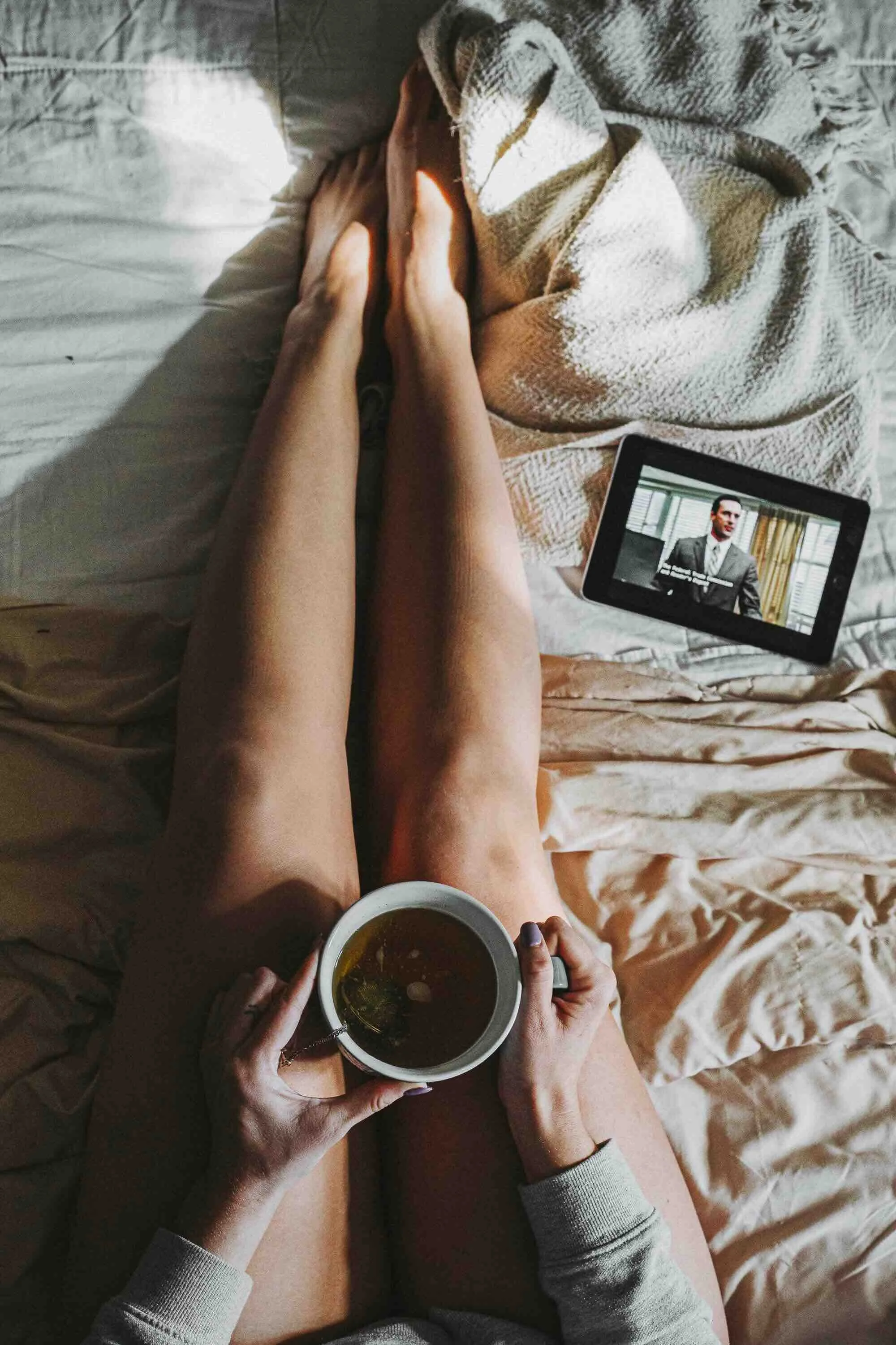 Personne allongée au lit avec les jambes croisées, tenant une tasse de thé, avec une tablette à côté montrant un homme en costume, et une couverture enroulée partiellement autour d'elle.
