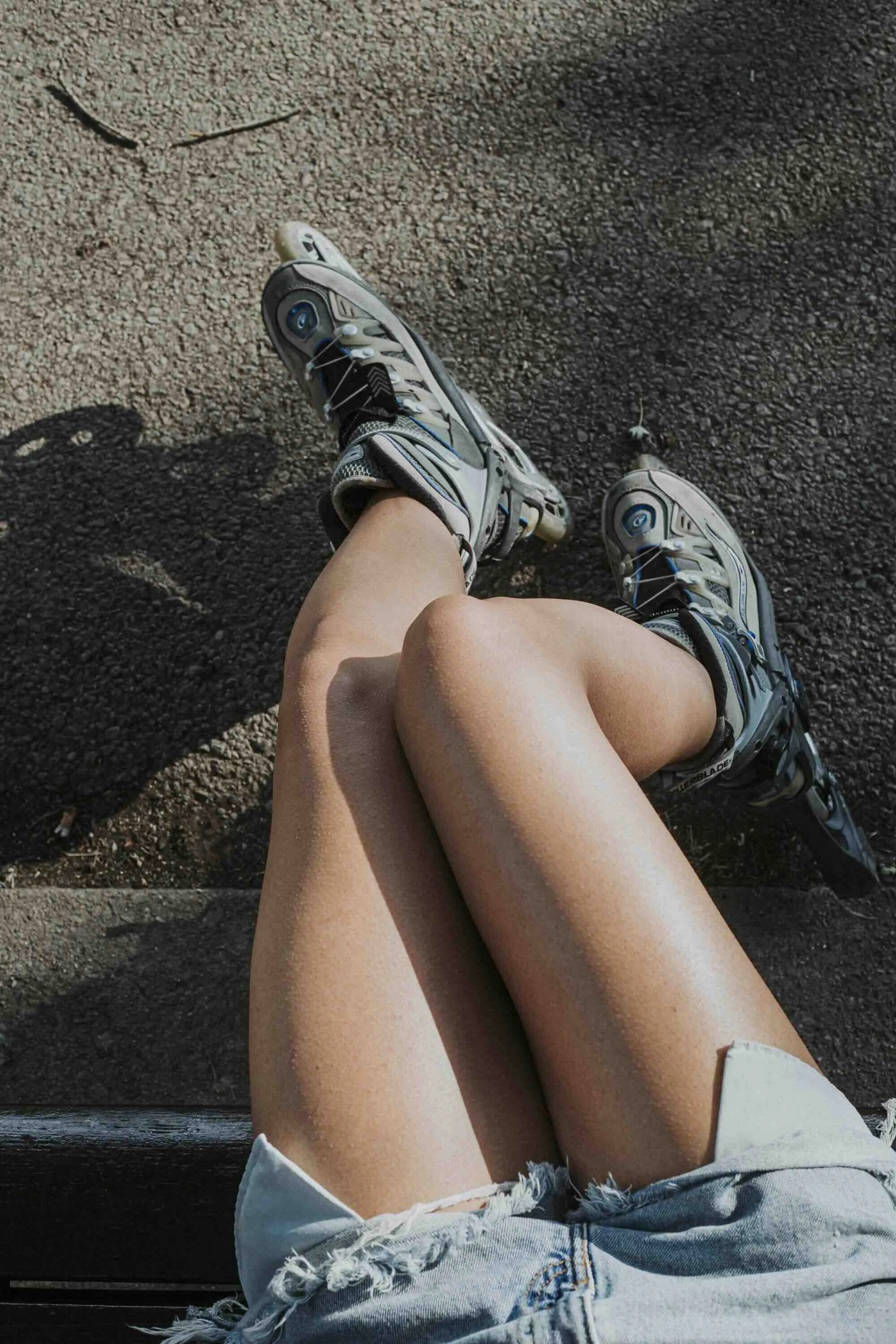Jambes de personne en shorts déchirés, portant des rollers, assise sur un banc, avec le sol en asphalte en dessous.