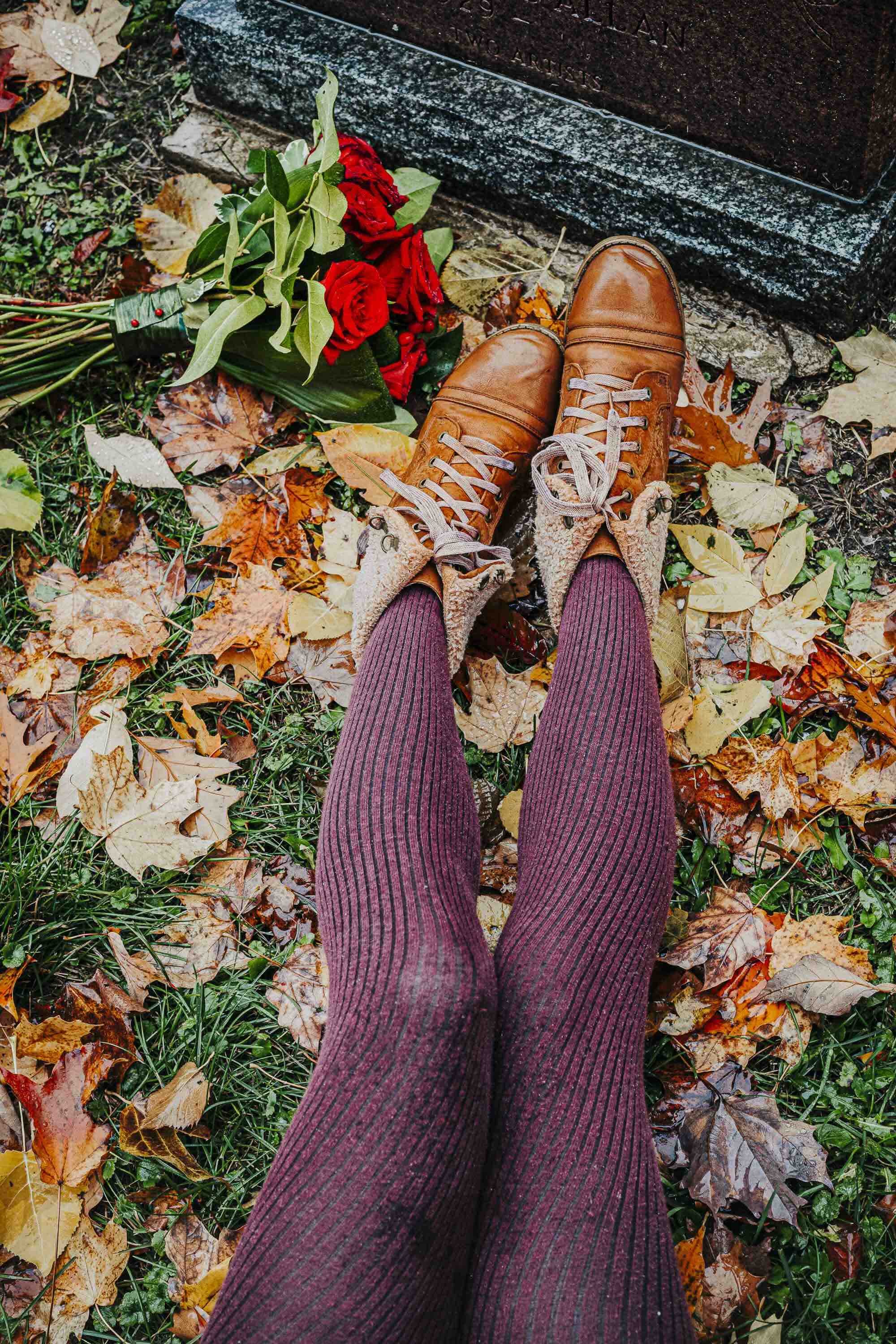 Pieds portant des bottes en cuir marron, habillés de chaussettes épaisses de couleur bordeaux, sur un sol recouvert de feuilles mortes.