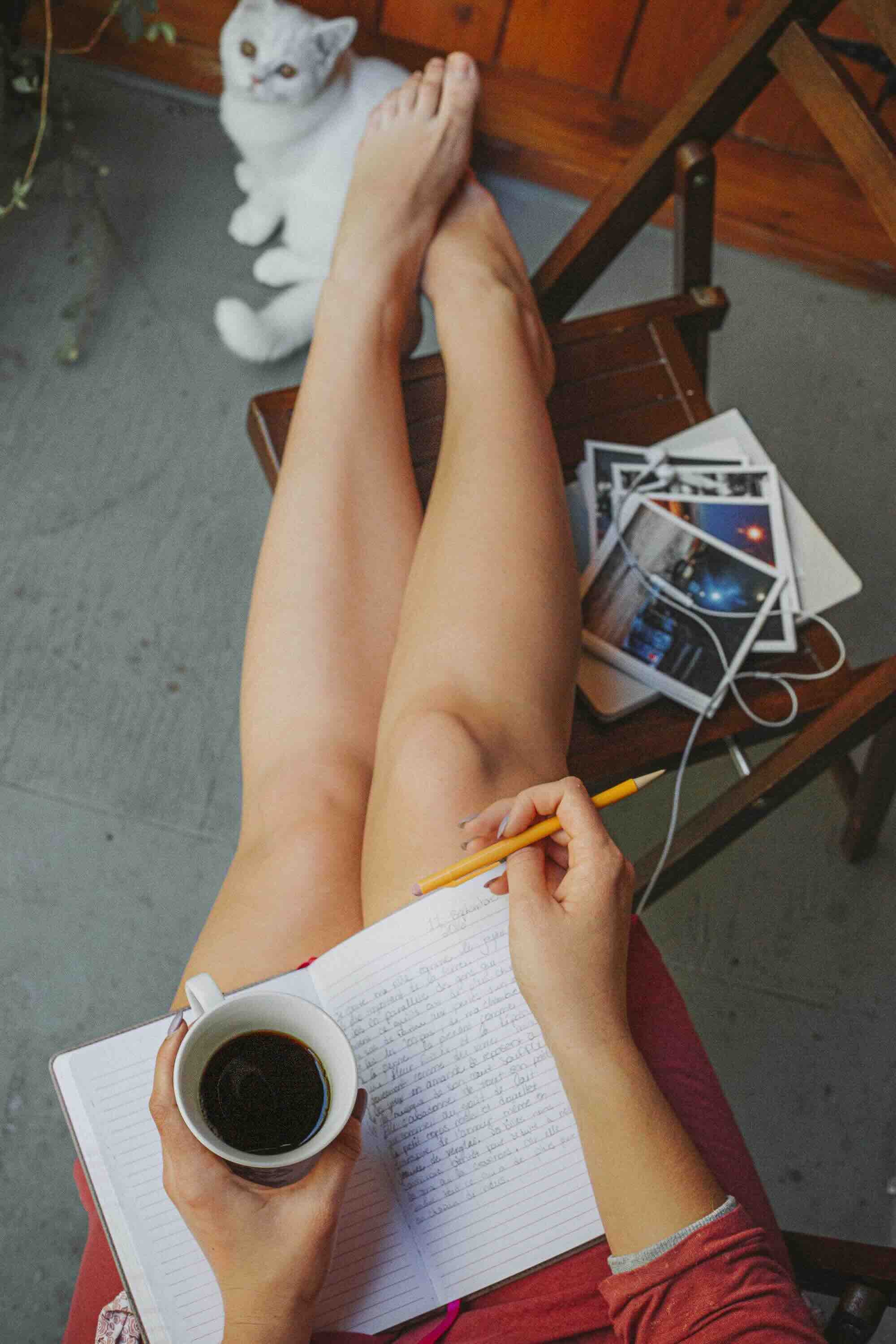 Une personne assise, tenant un carnet de notes avec un stylo, une tasse de café dans une main, et des photos empilées sur une chaise à côté. Un chat blanc assis à proximité regarde en direction de la personne.