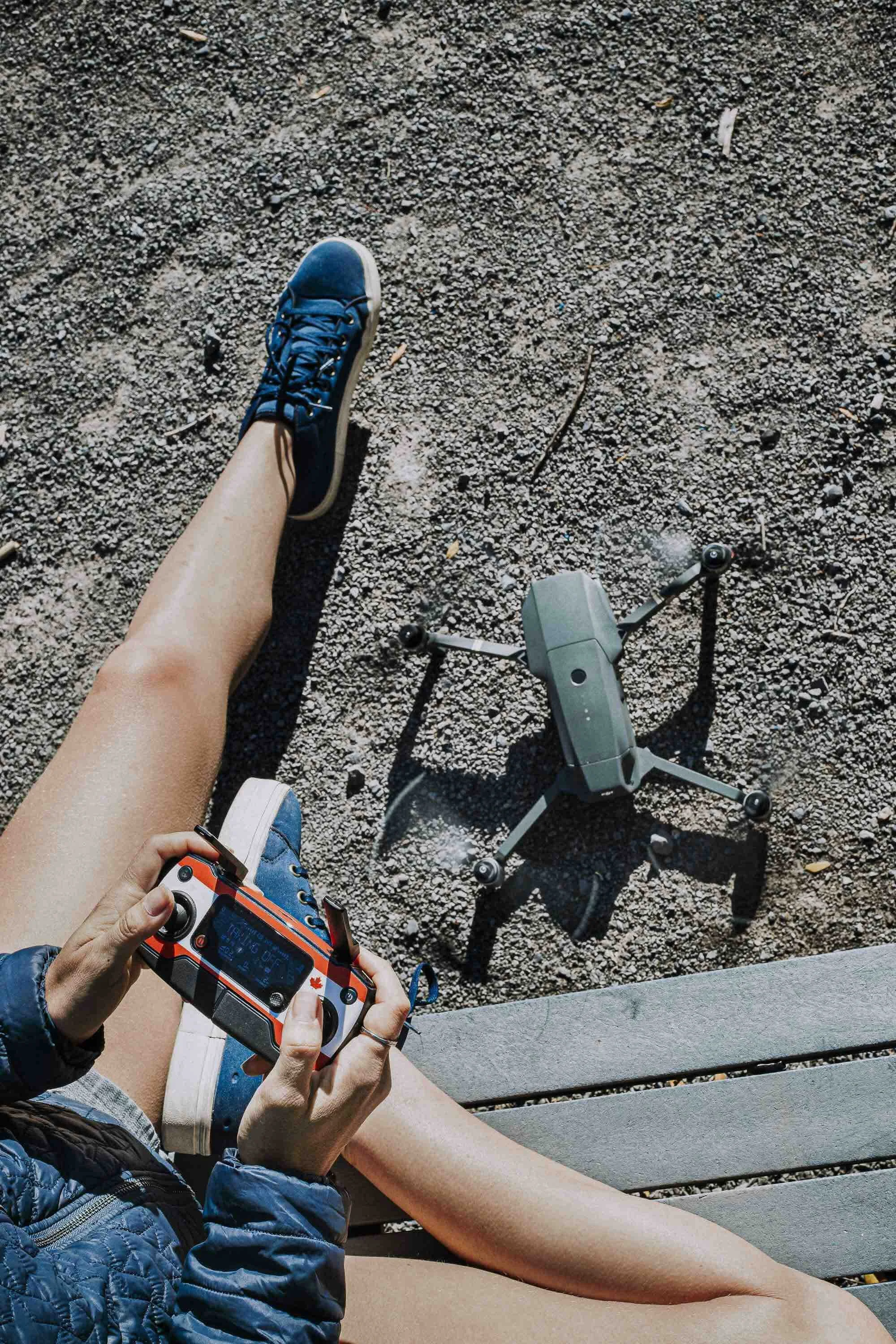 Une personne assise sur un banc, tenant une télécommande ou un contrôleur de drone, avec un drone au sol à côté sur un sol en gravier.
