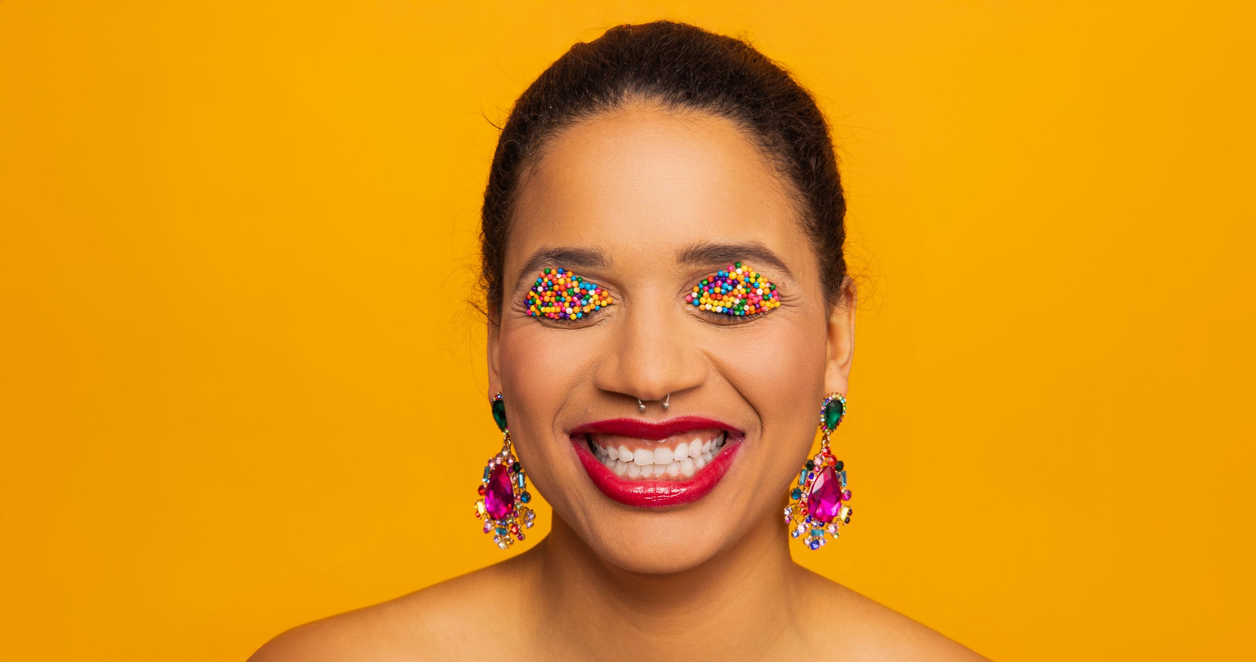 Une femme souriante avec des yeux couverts de paillettes multicolores et portant de grosses boucles d'oreilles scintillantes, devant un fond jaune