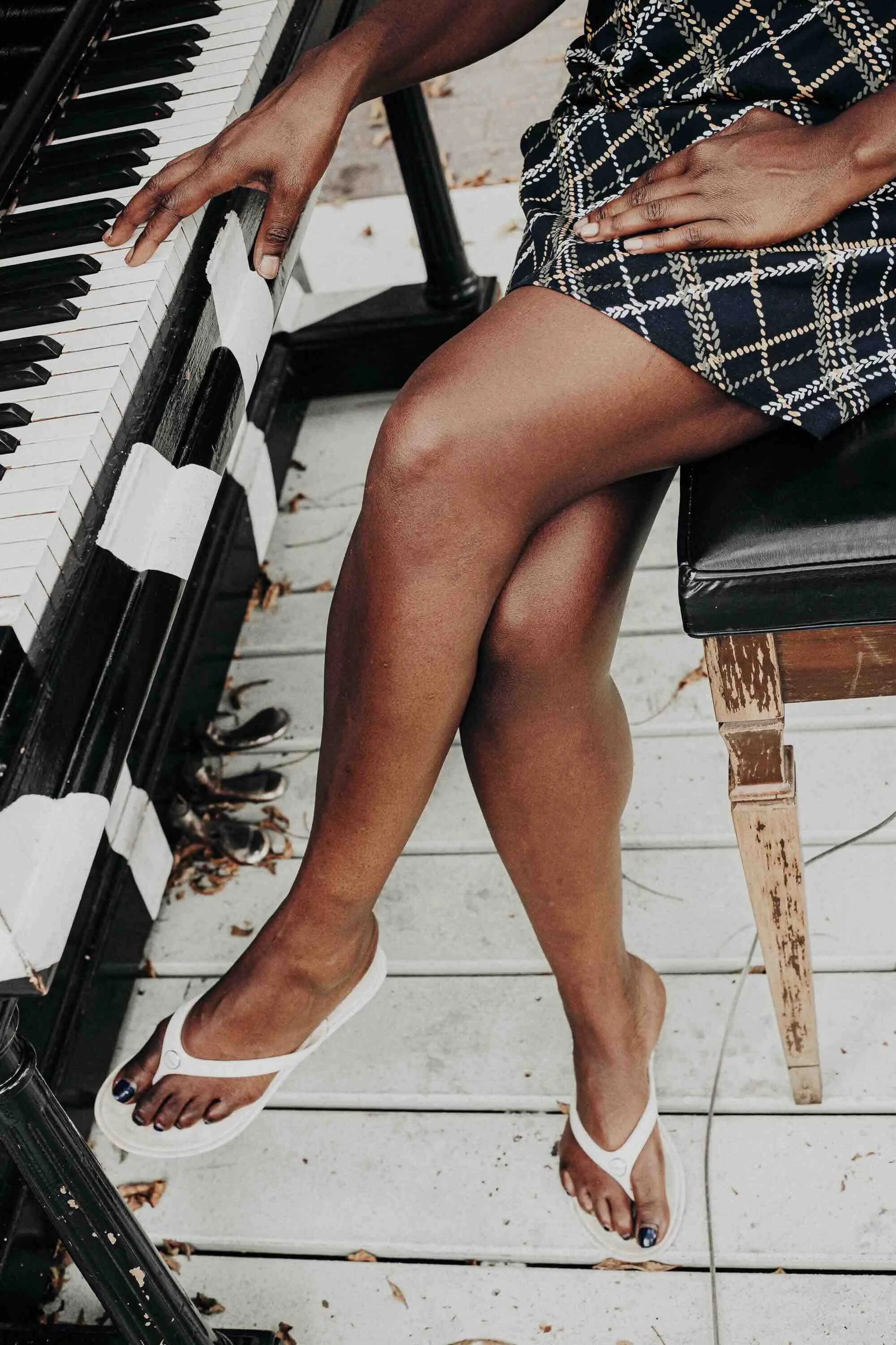 Une personne jouant du piano en extérieur, assise sur un banc en bois. Elle porte une robe noire à motif de lignes de chaîne et des sandales blanches. La photo montre ses jambes croisées et une main sur le clavier du piano.