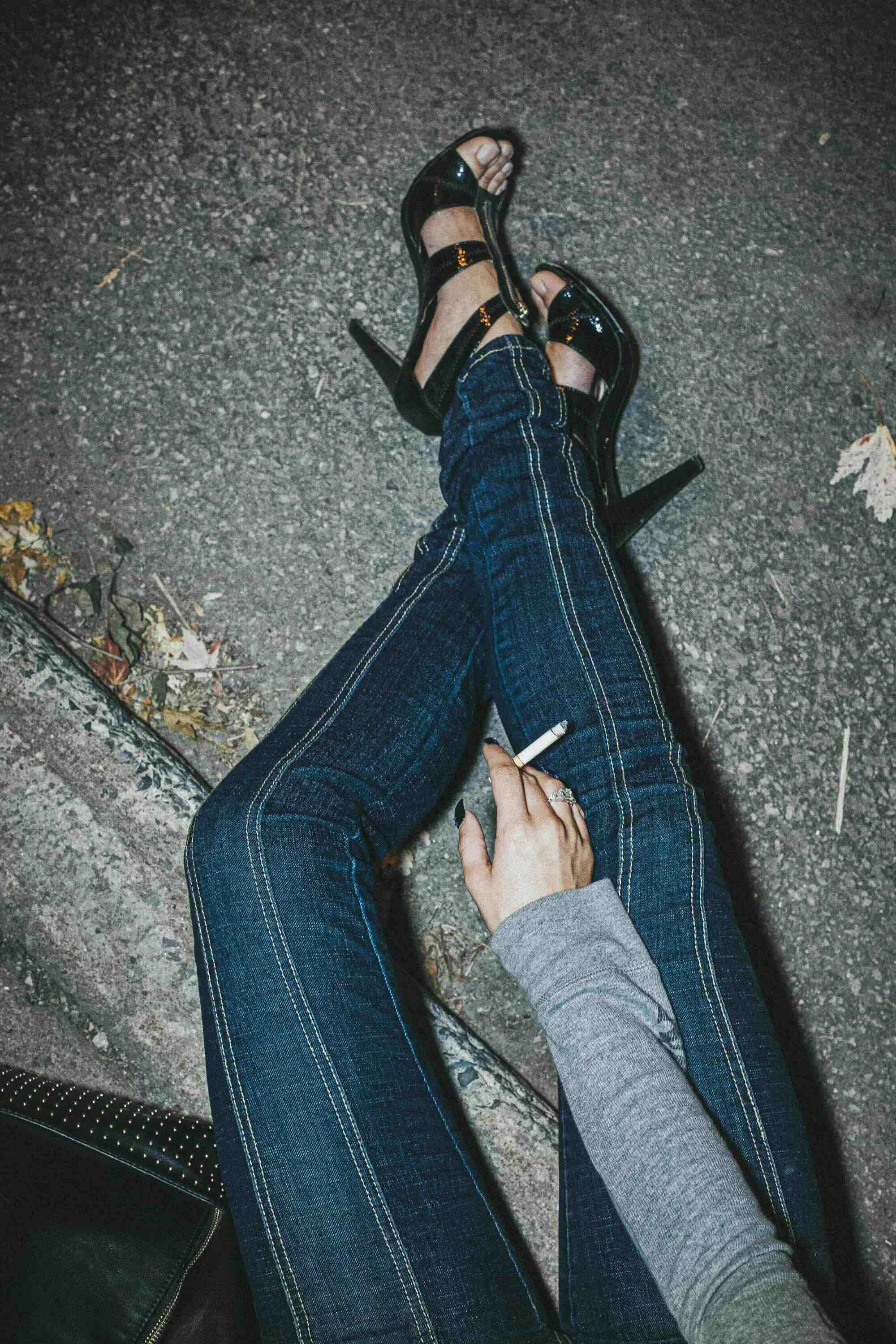 Personne allongée dans la rue portant des sandales à talons hauts, jeans et un sweat gris, tenant une cigarette en main.