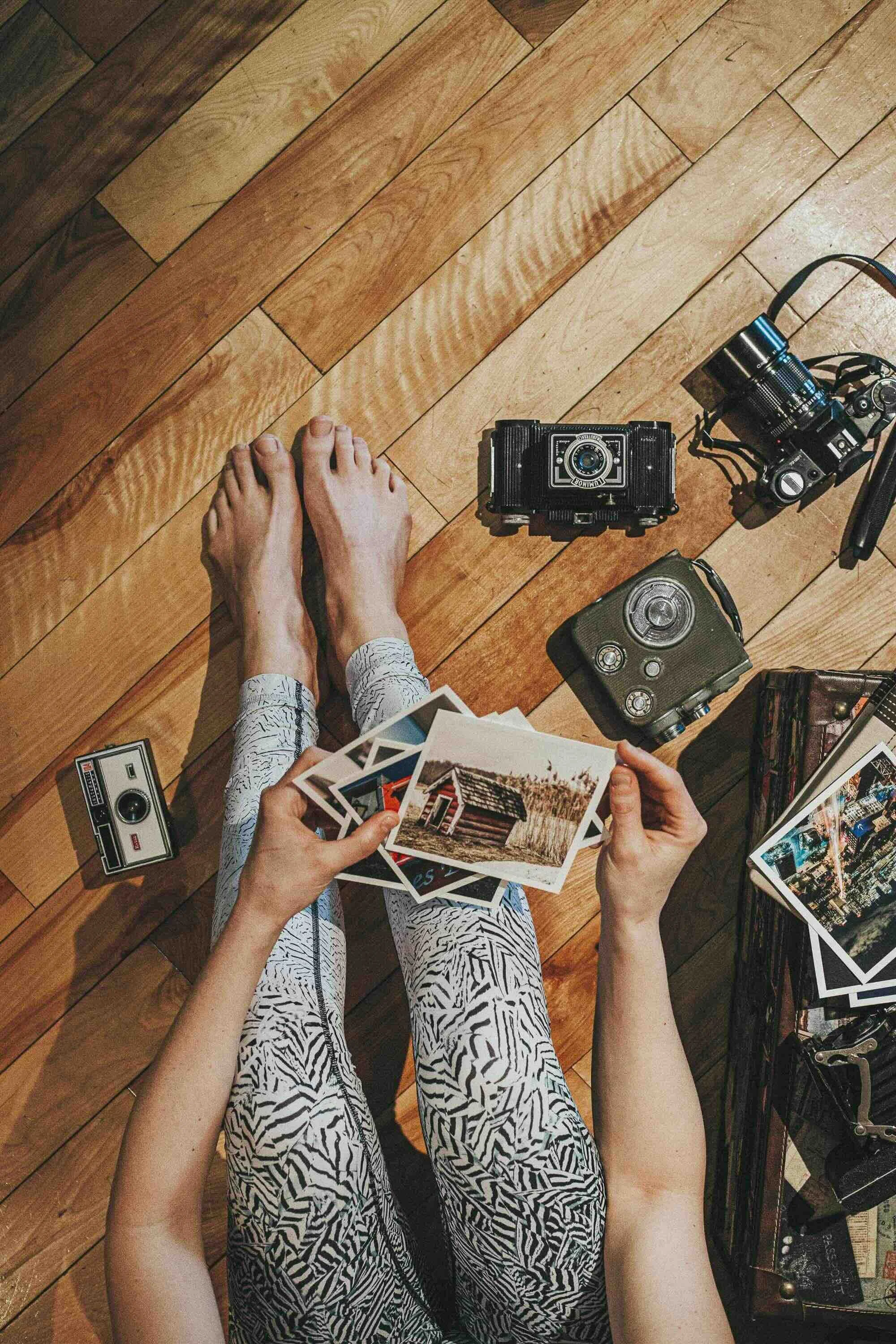 Une personne assise par terre en train de feuilleter des photos, entourée de plusieurs appareils photo vintage et d'une valise contenant des photos. La vue est d'en haut.