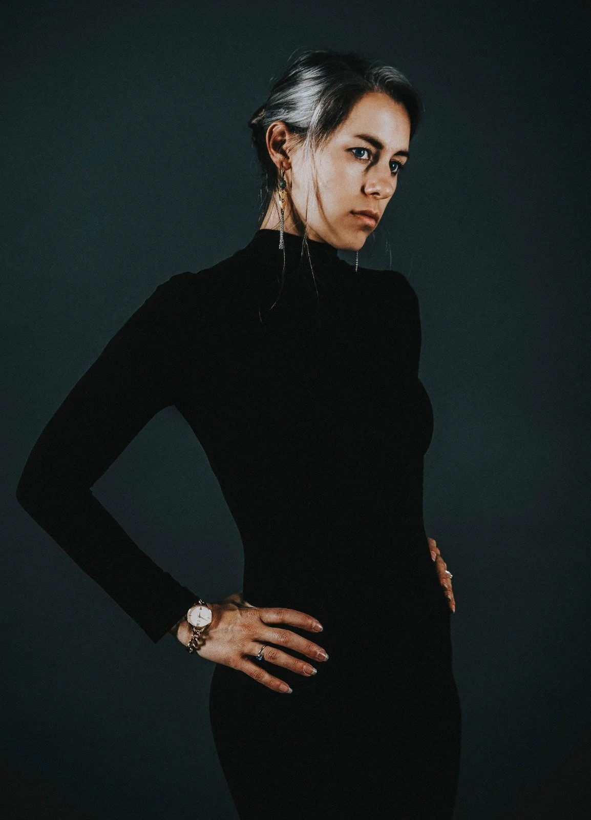 Une femme habillée en noir, avec des cheveux gris mêlés de noir, pose avec les mains sur les hanches contre un fond sombre.