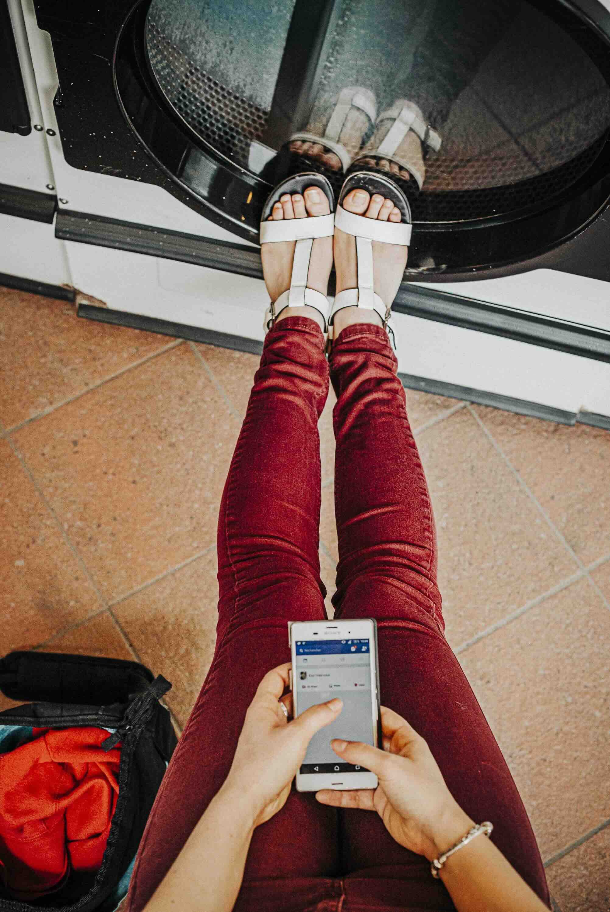 Une personne en jeans rouges portant des sandales blanches, assise près d'une machine à laver, tenant un téléphone portable avec une main, avec un sac orange-vert à côté.