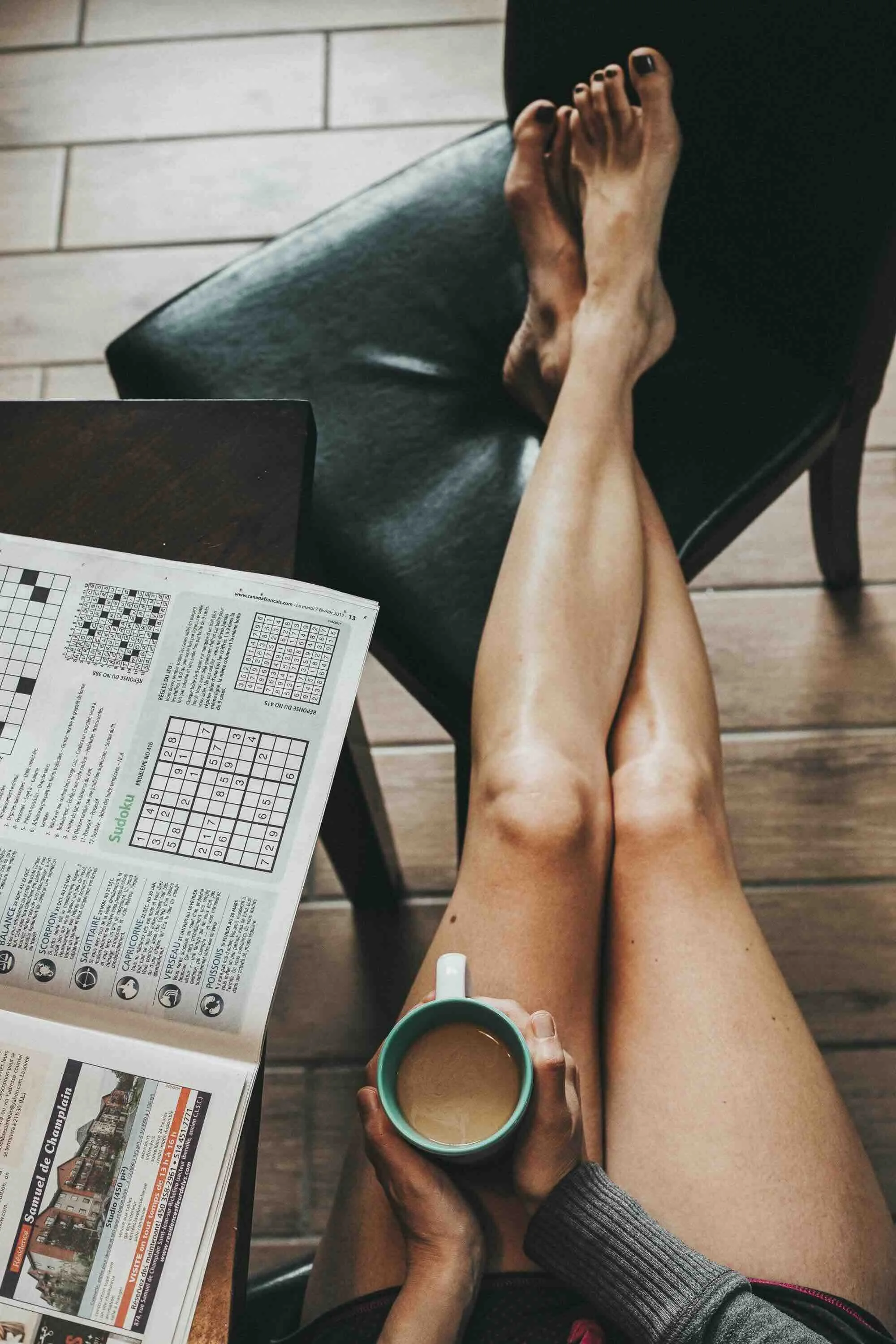 Une personne assise sur une chaise noire, jambes croisées, tenant une tasse de café. Sur la table en bois à côté, il y a un journal ouvert avec des sudoku.