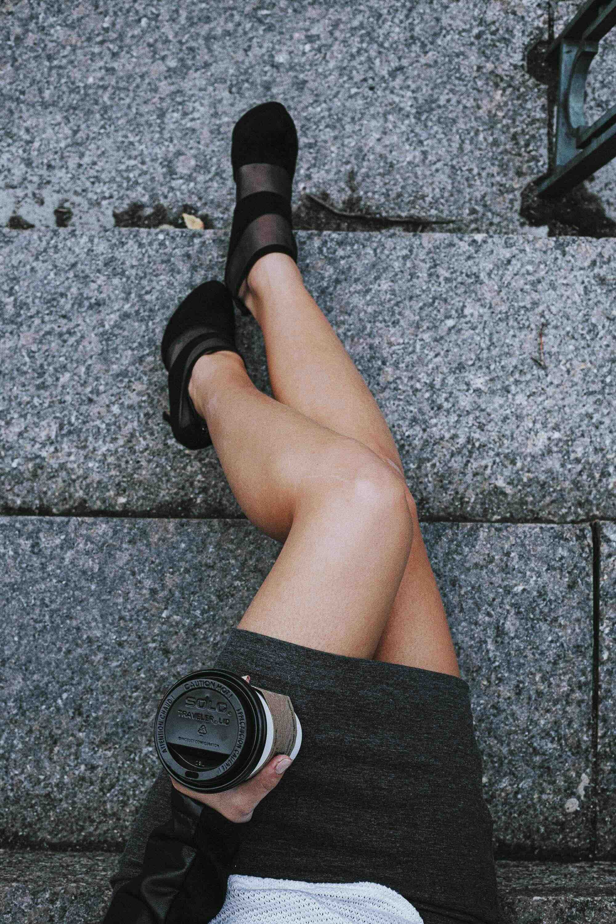 Jambes croisées d'une personne assise sur un trottoir en granite, portant des chaussures noires, un short noir, et tenant une tasse à café