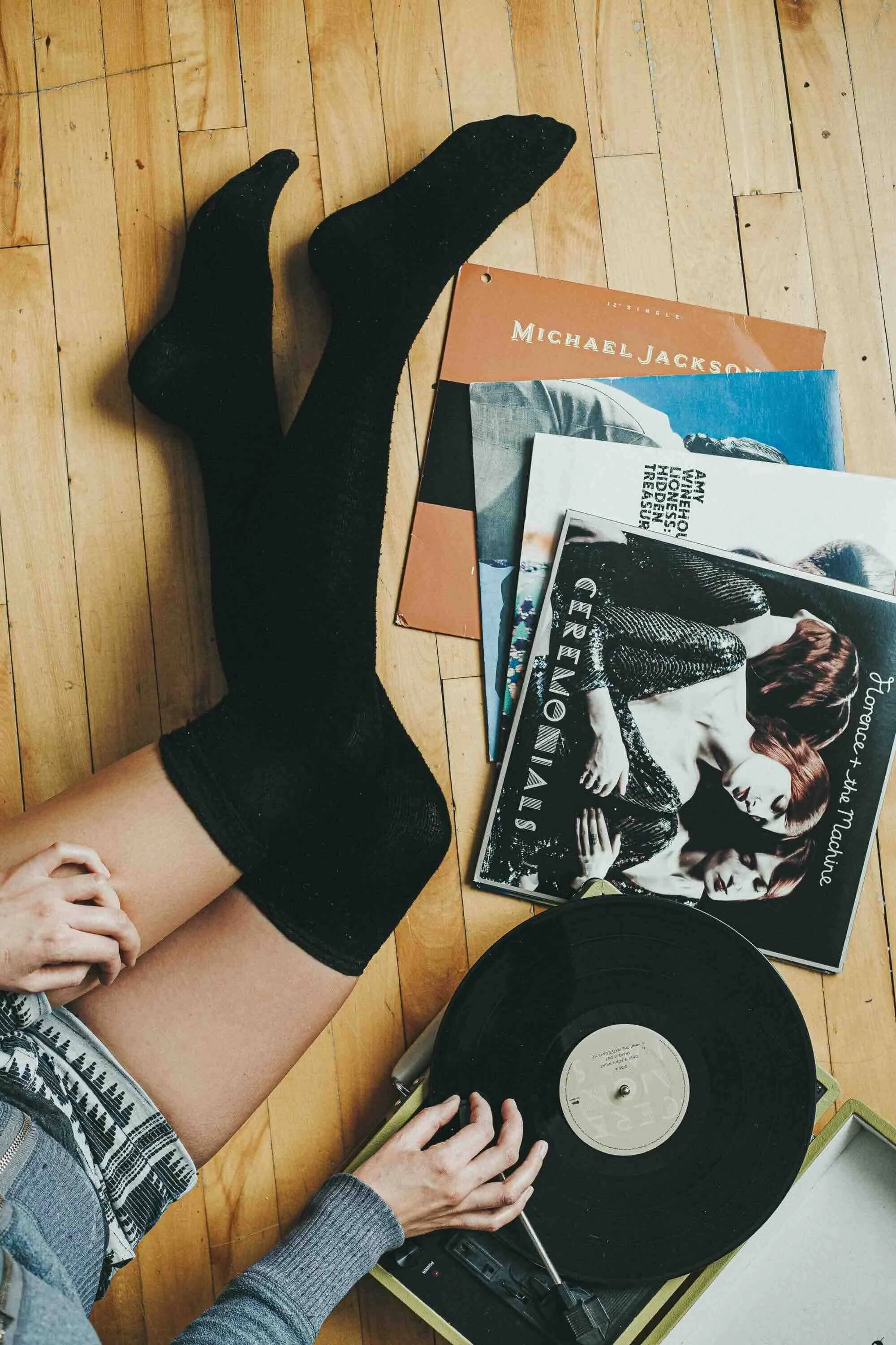 Une personne assise sur un parquet en train de manipuler un tourne-disque avec un disque vinyle. À côté, il y a une pile de magazines, parmi lesquels on peut reconnaître un magazine de Greta Van Fleet et un autre de Michael Jackson.