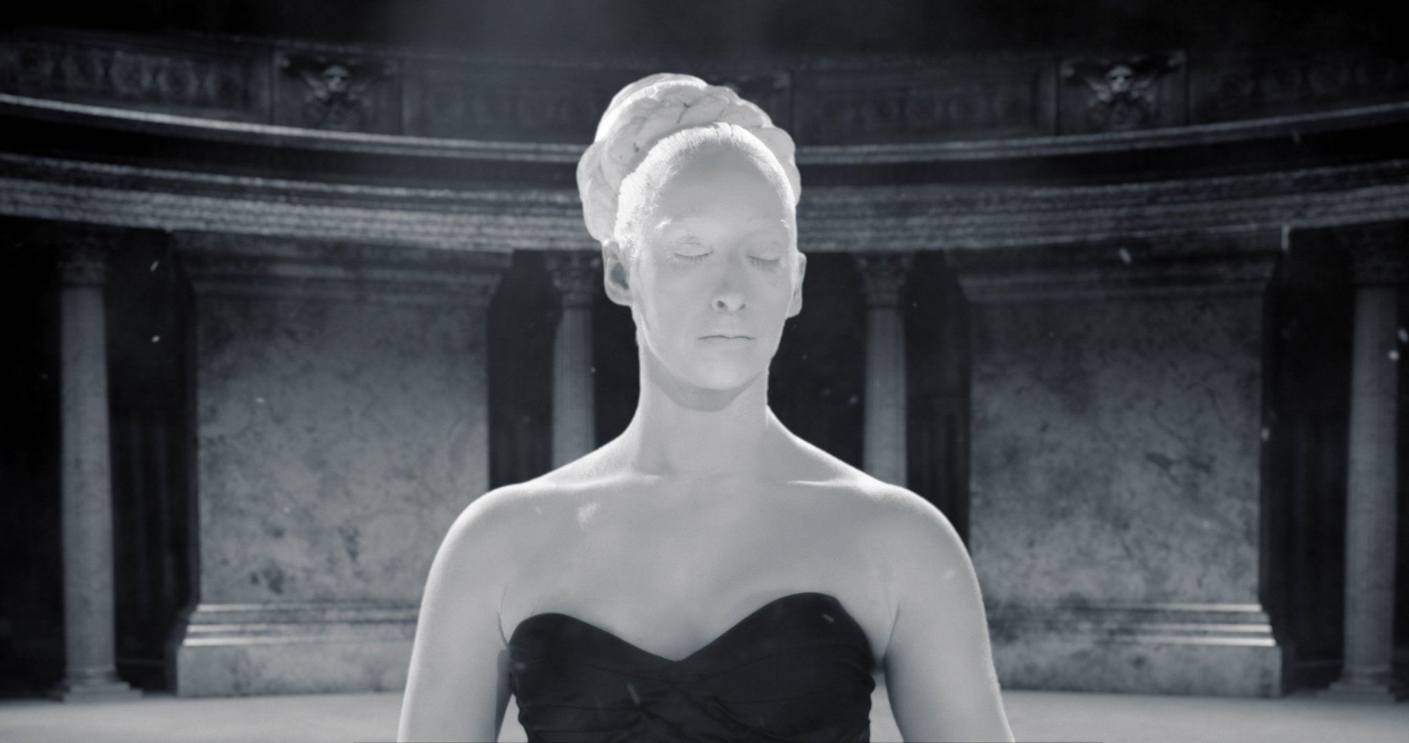 Une femme en robe noire, yeux fermés, dans un environnement monochrome, avec une architecture ancienne en arrière-plan.