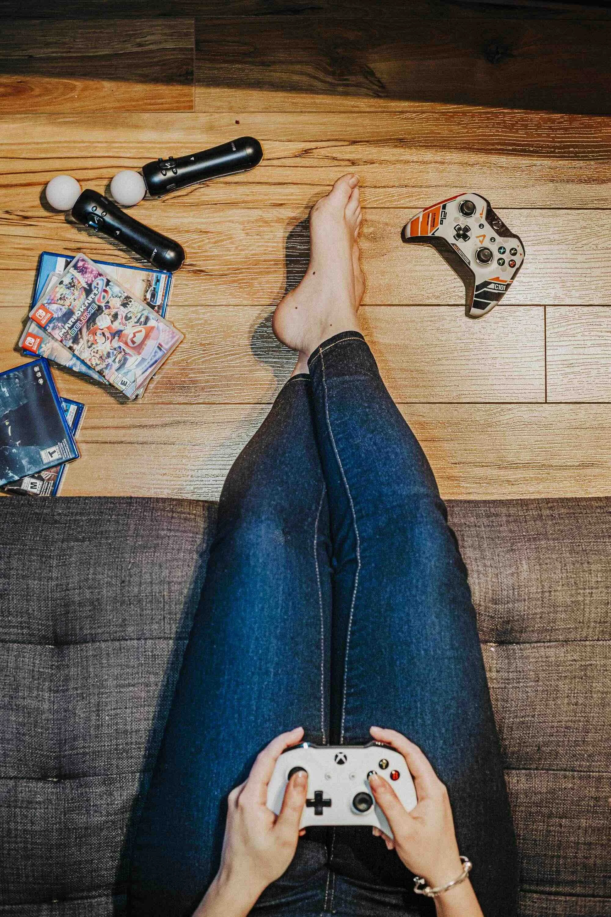 Personne assise sur un canapé avec des jambes croisées, tenant une manette Xbox, avec une console de jeux, des jeux vidéo, un autre contrôleur, et un appareil de massage sur le sol en bois devant.