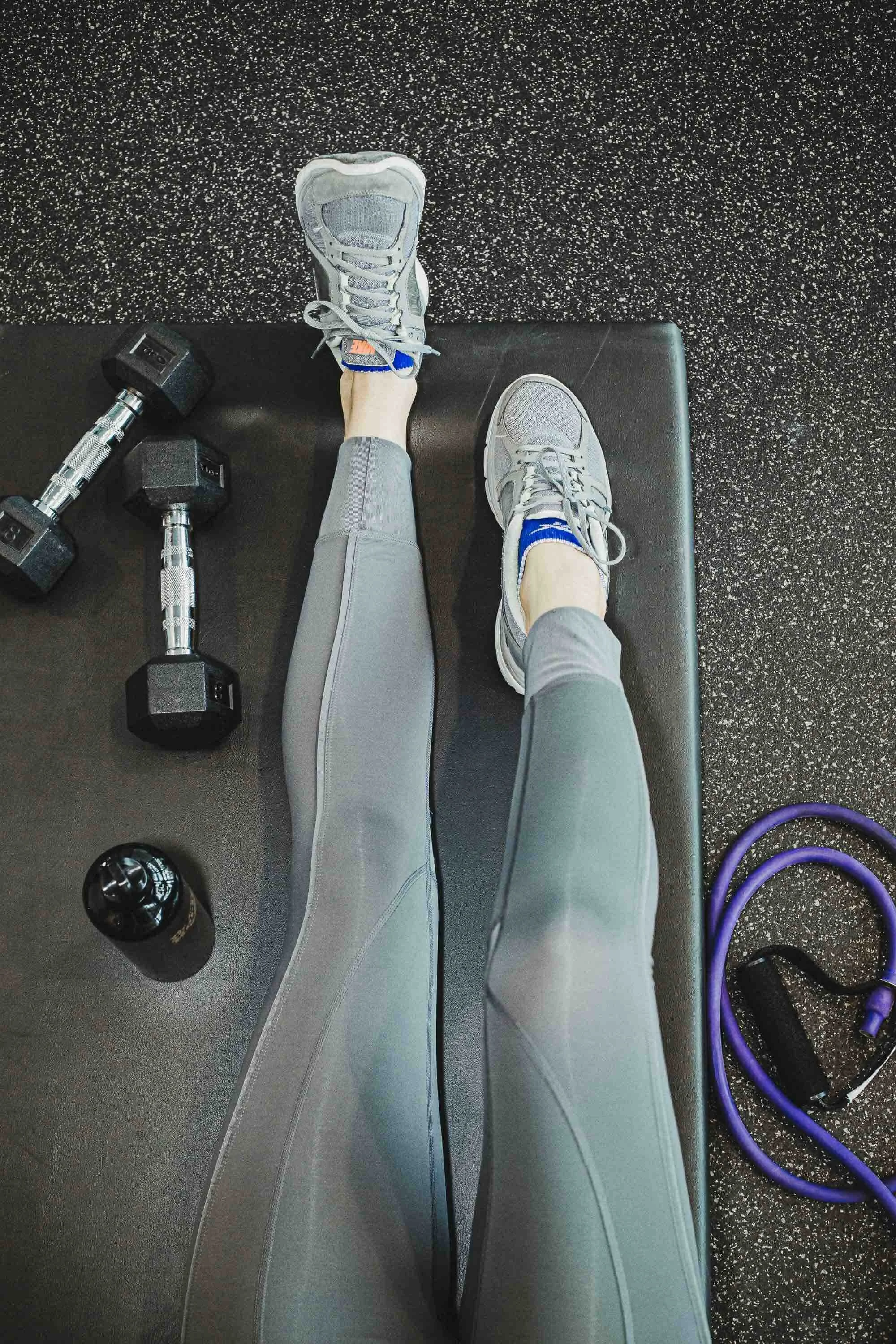 Jambes d'une personne en leggings gris, faisant de l'exercice avec des haltères, une bouteille d'eau et une bande de résistance à proximité dans une salle de gym.