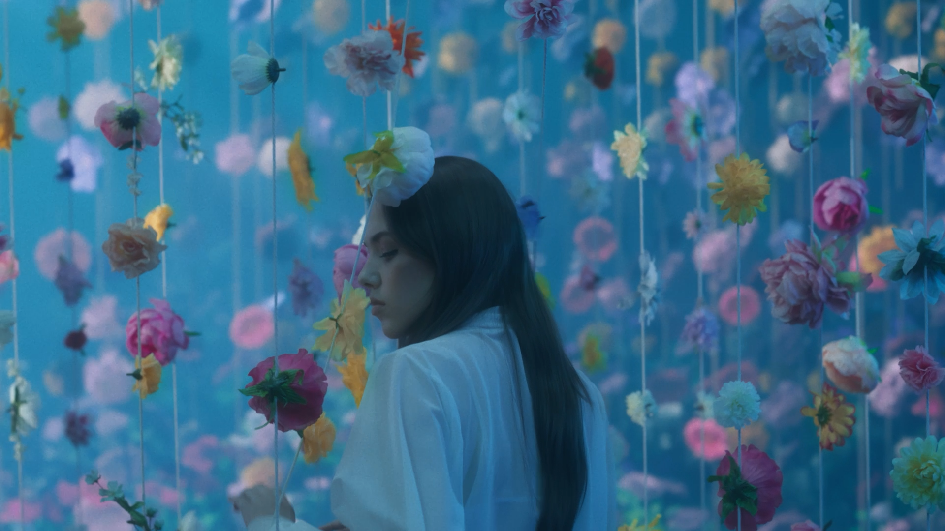 Jeune femme aux yeux fermés dans un décor avec des fleurs suspendues dans l'air, ambiance douce et mystérieuse.