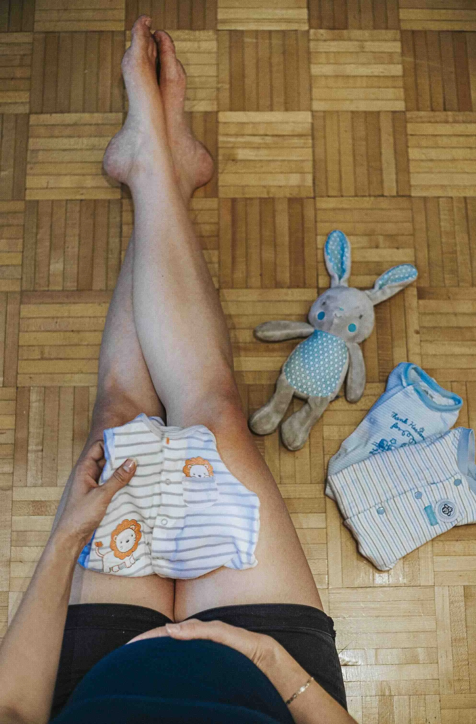Une personne assise, en train de changer un enfant, avec des vêtements de bébé et une peluche lapin à côté sur un sol en parquet.