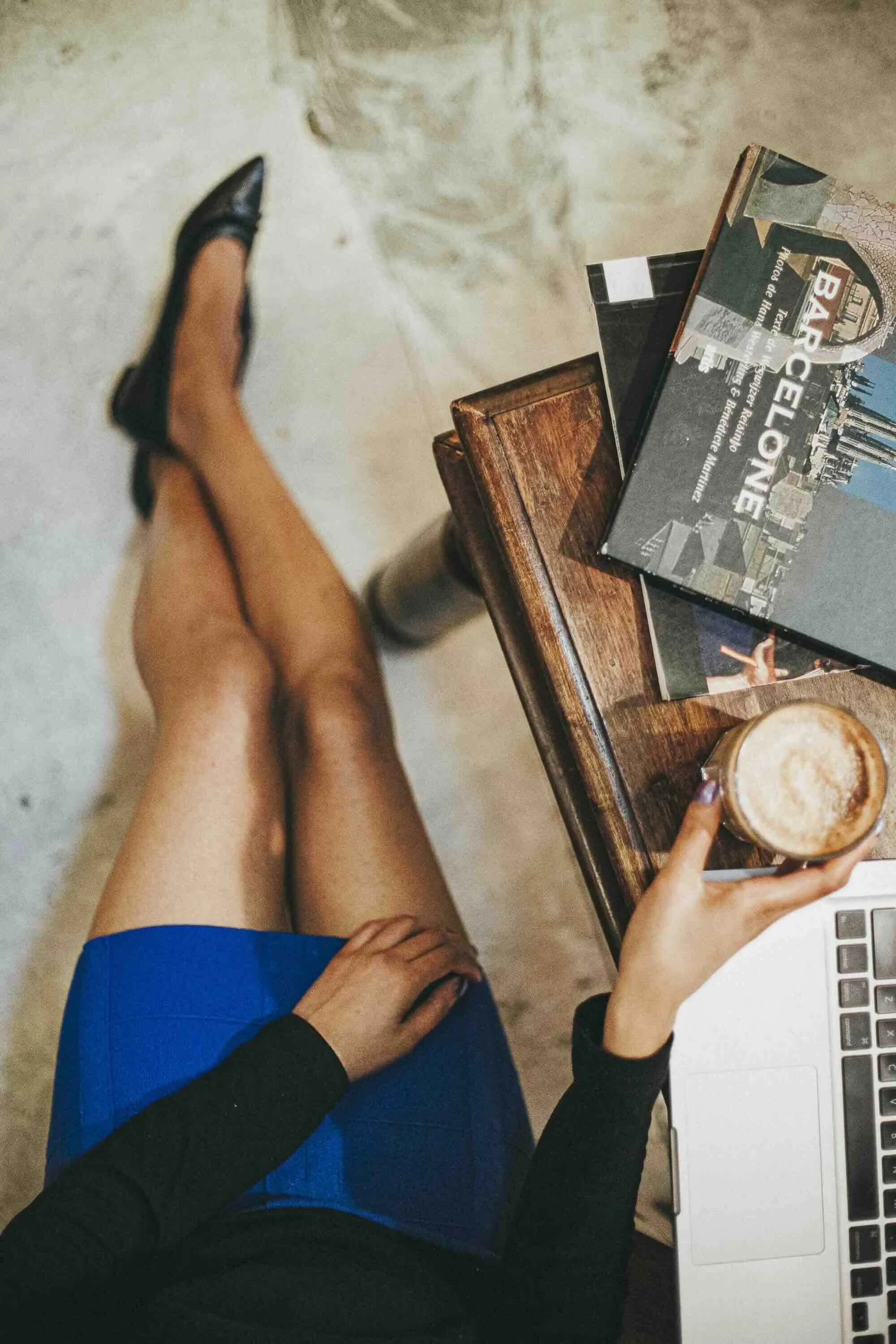 Une personne assise, portant une jupe bleue, tient une tasse de café et un ordinateur portable sur une table en bois, avec des magazines posés dessus.