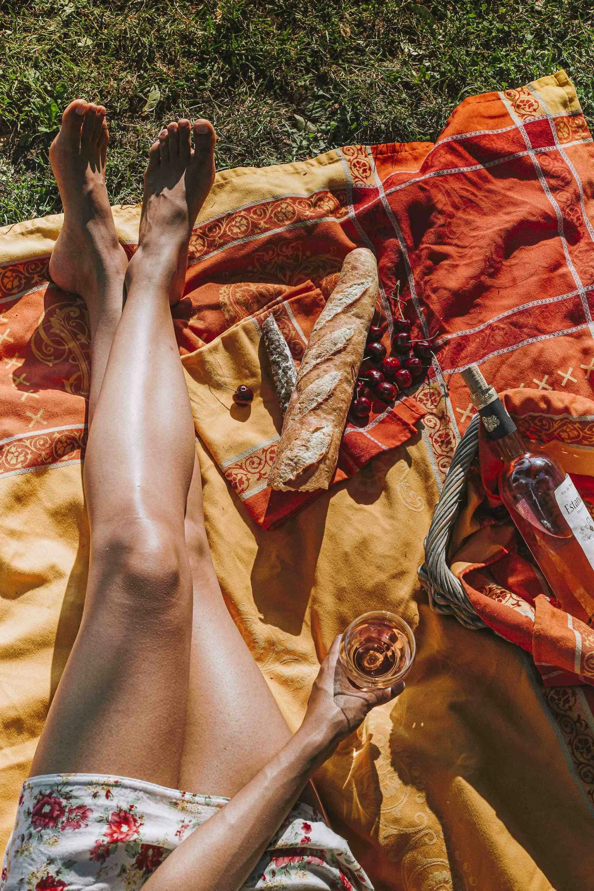 Une personne allongée sur une nappe orange en extérieur, tenant un verre de vin rosé. Devant elle, un pique-nique avec du pain, des cerises, une bouteille de vin rosé et un couteau. L'herbe verte entoure la scène.
