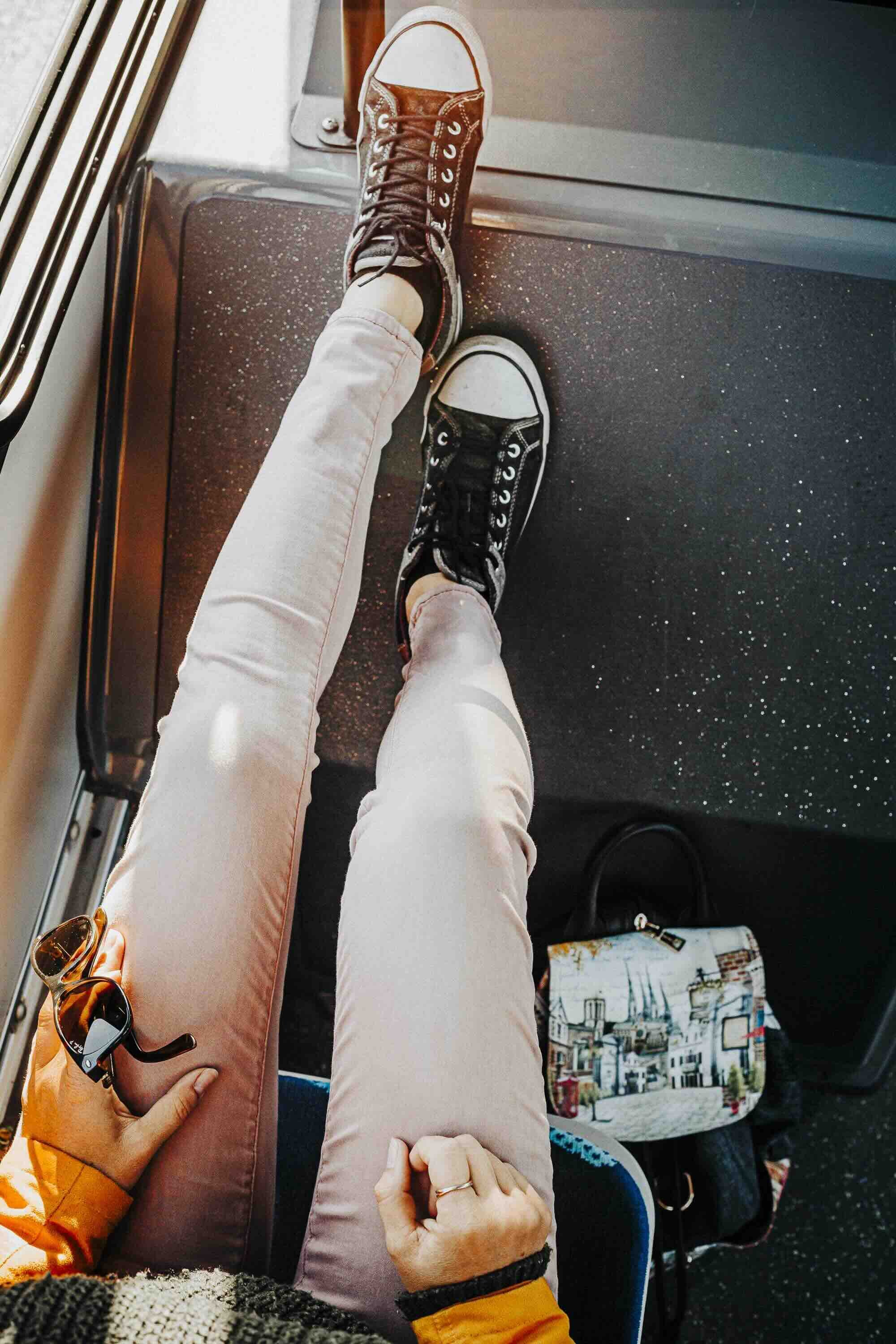 Les jambes d'une personne assise dans un avion, portant des baskets Converse noires et blanches, des pantalons roses pâles, avec un sac à main aux motifs de bâtiments et de ville en bas à côté, et des lunettes de soleil dans la main gauche.