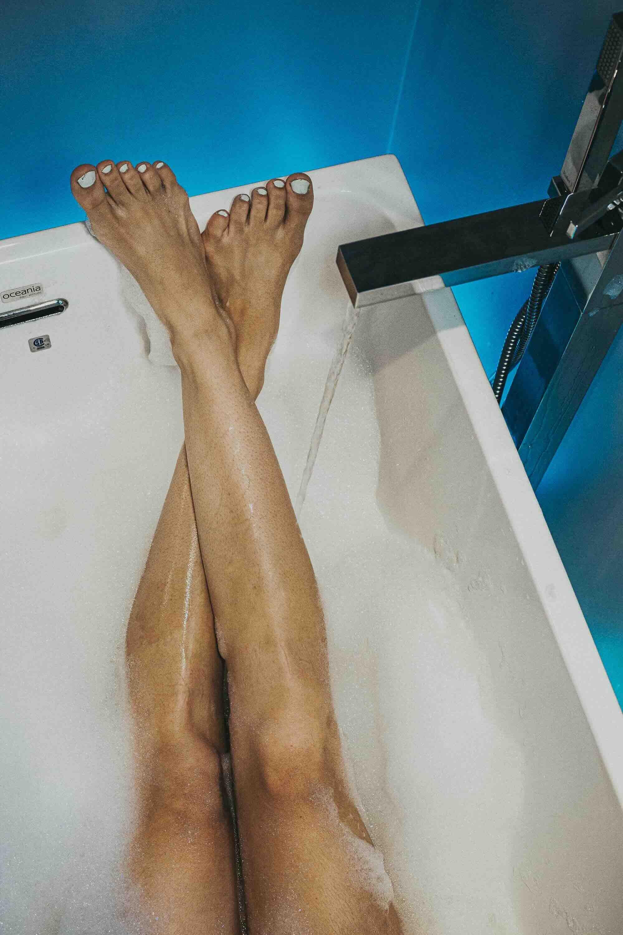 Jambes d'une personne prenant un bain dans une baignoire en eau, avec de la mousse sur la peau, sous une douche miouverte.