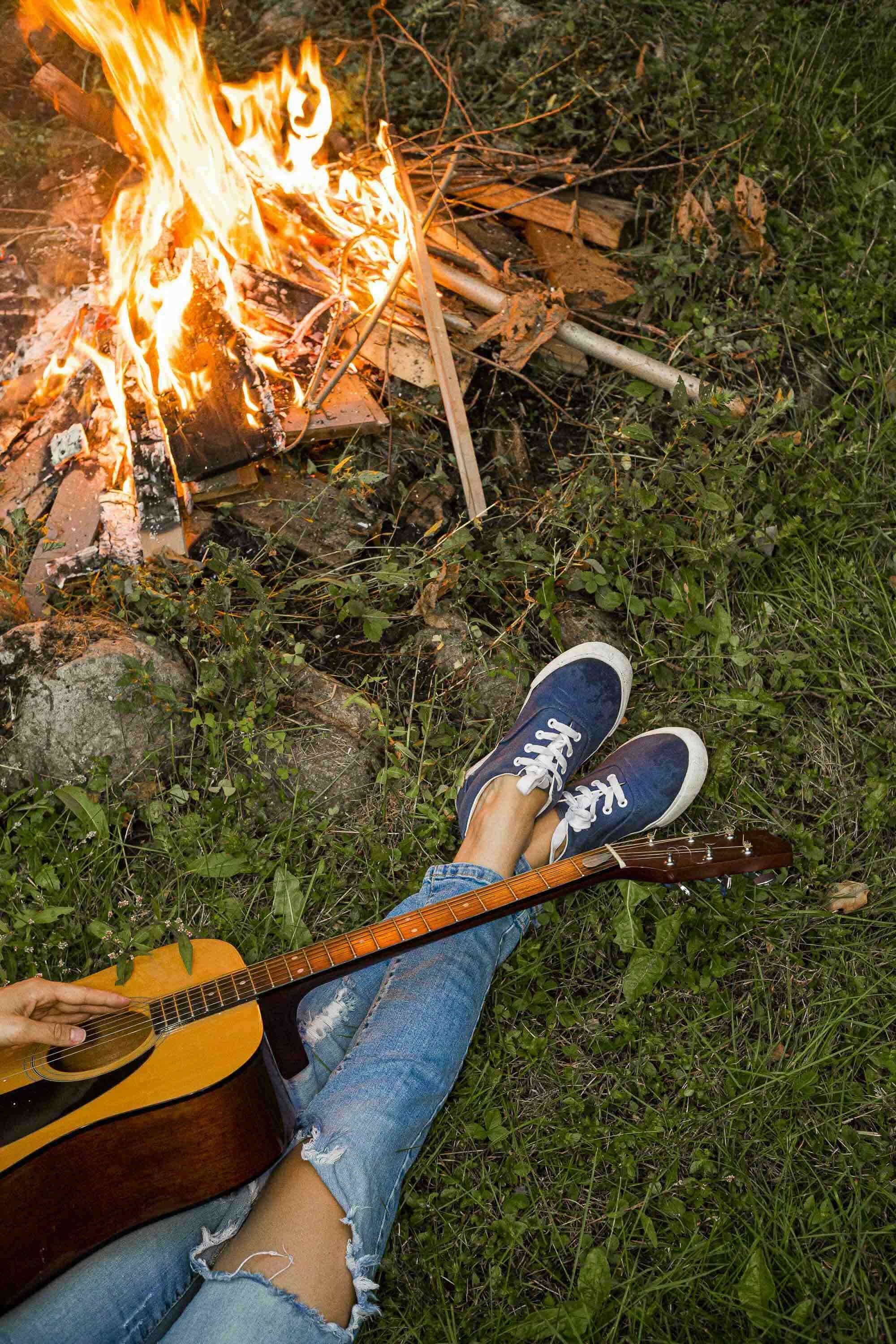 Personne assise sur l'herbe, jouant de la guitare près d'un feu de camp, portant des jeans déchirés et des chaussures bleues.