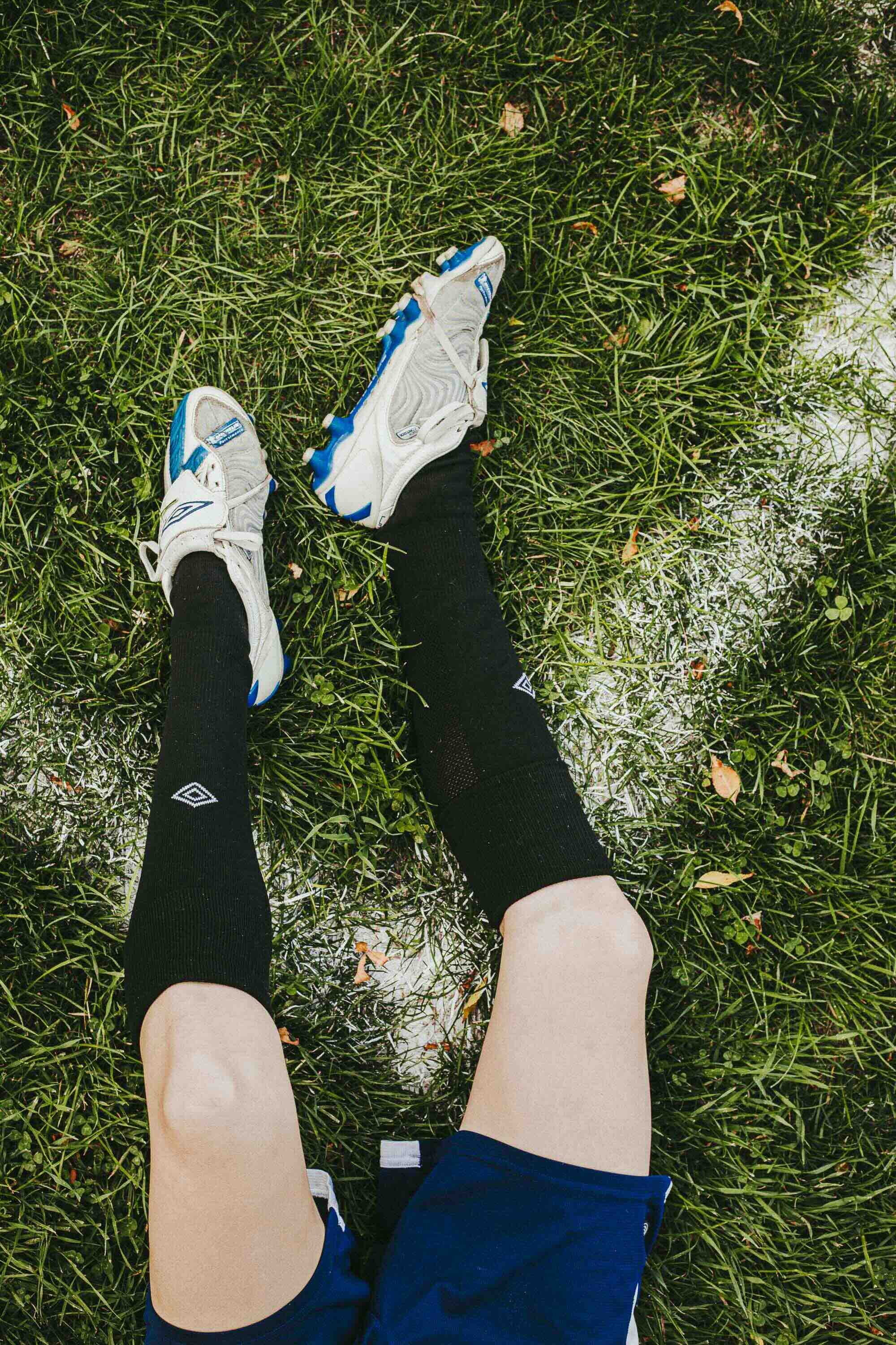 Jambes d'une personne en short, portant des chaussettes longues noires, des chaussures de sport beige et bleu, allongée sur de l'herbe avec quelques feuilles tombées.