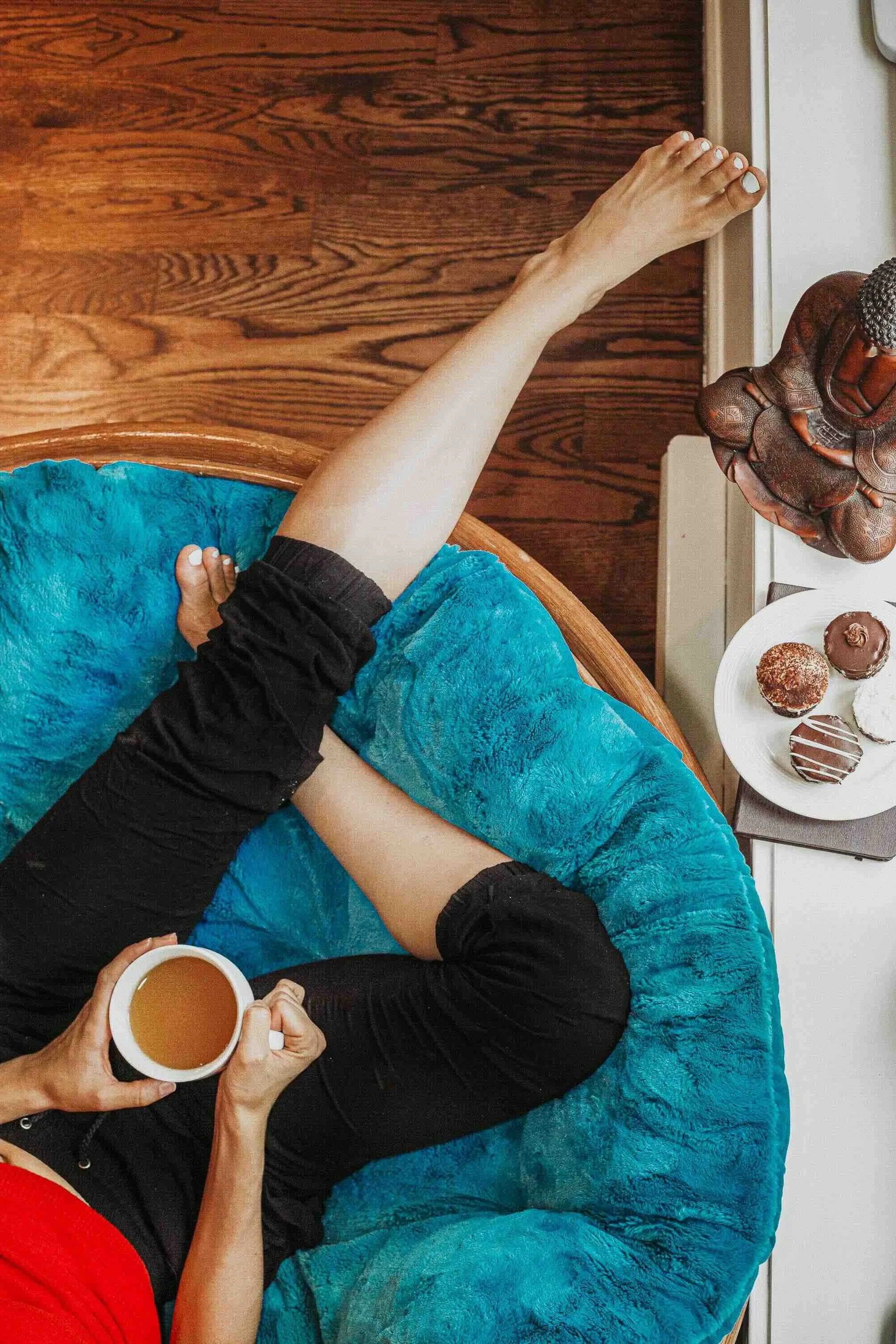 Une personne assise dans un fauteuil bleu, tenant une tasse de café ou thé, avec un plateau de pâtisseries sucrées sur une table à côté.