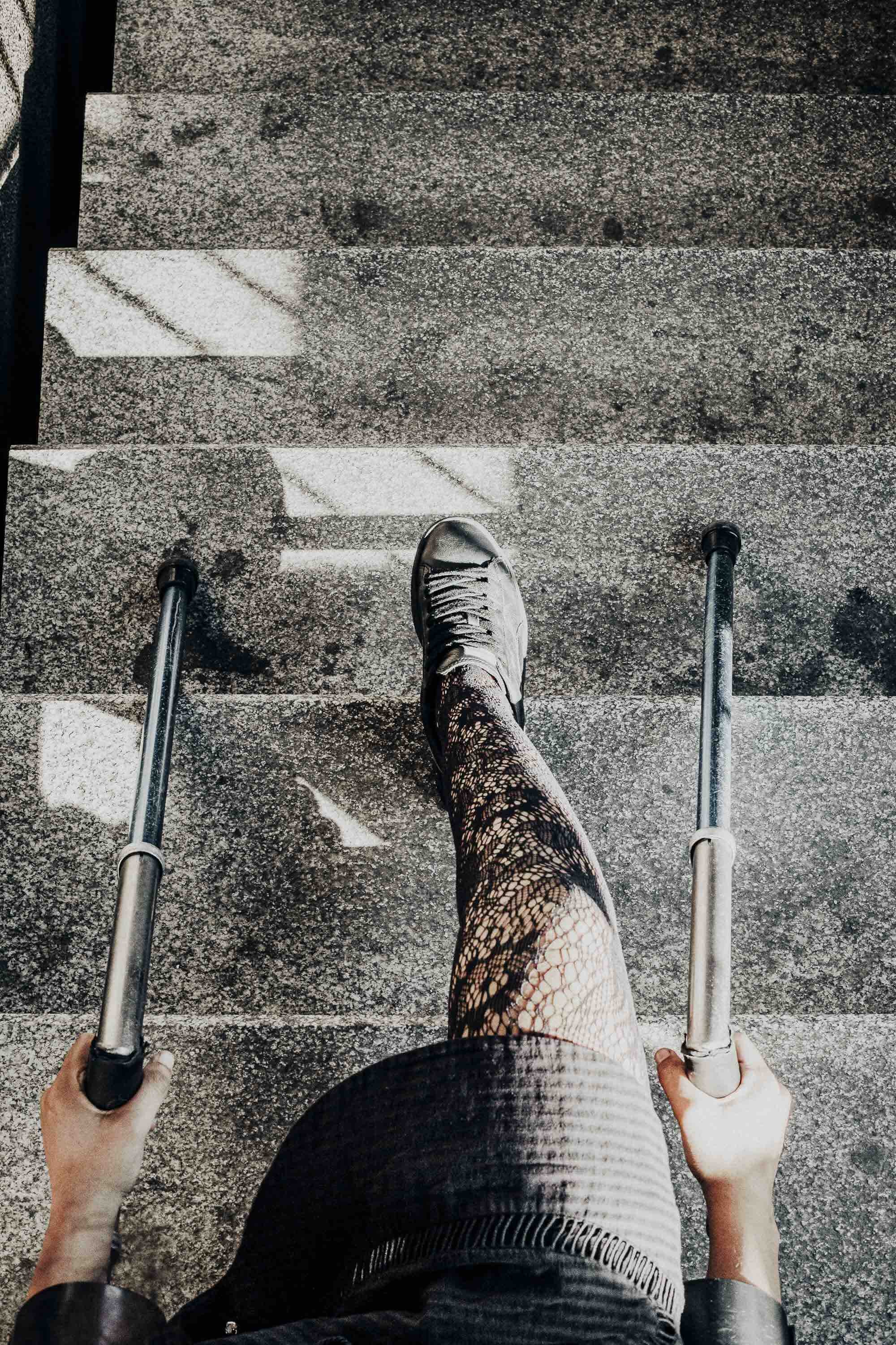Photo prise du haut d'une personne en fauteuil roulant avec des prothèses sur ses bras, montrant une jambe métallique dans des collants en dentelle, marchant sur un escalier en pierre.