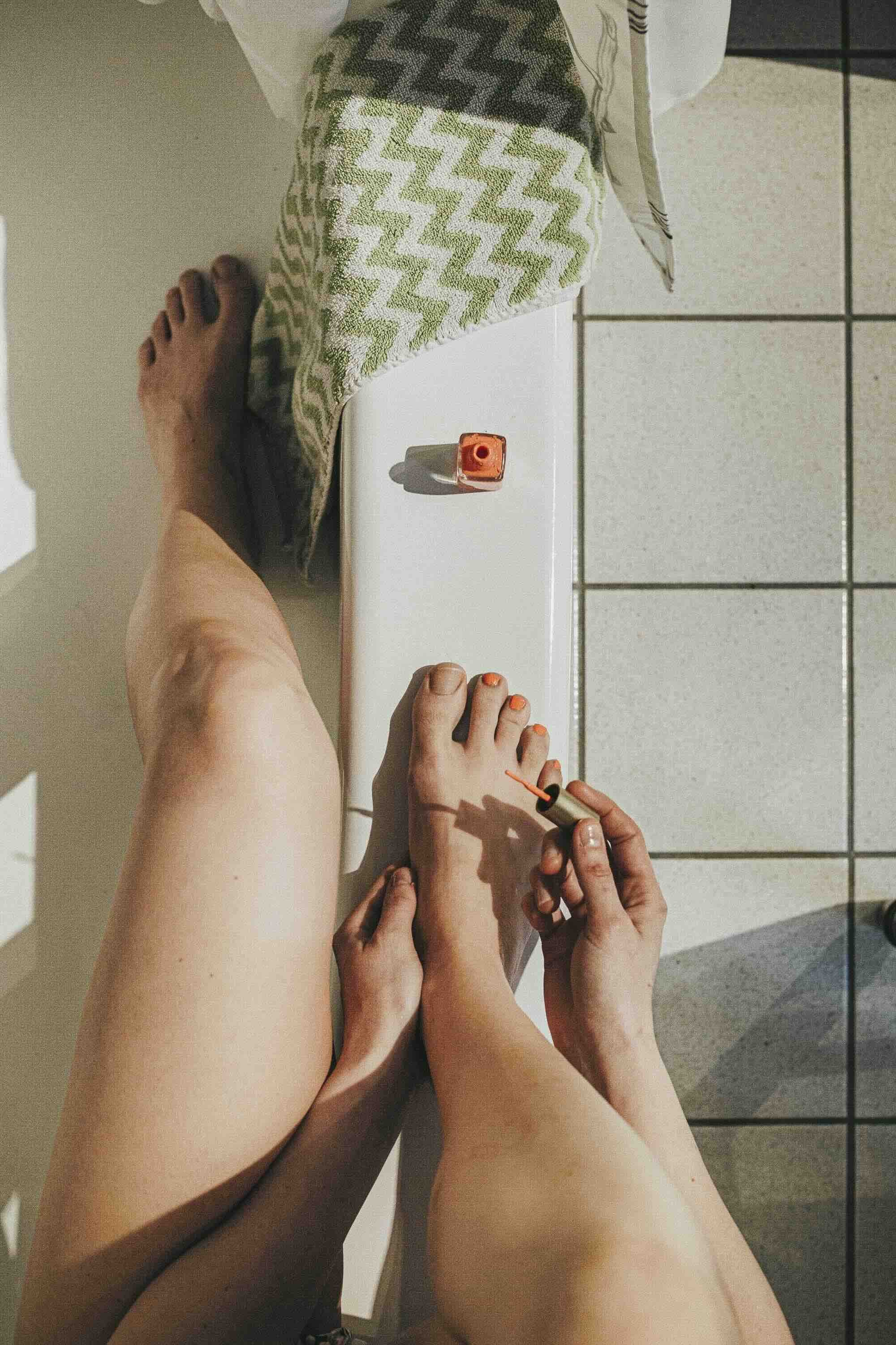 Une personne se peint les ongles de pieds avec du vernis orange, assise sur une surface blanche, avec une serviette et un fondu de la pièce en arrière-plan.