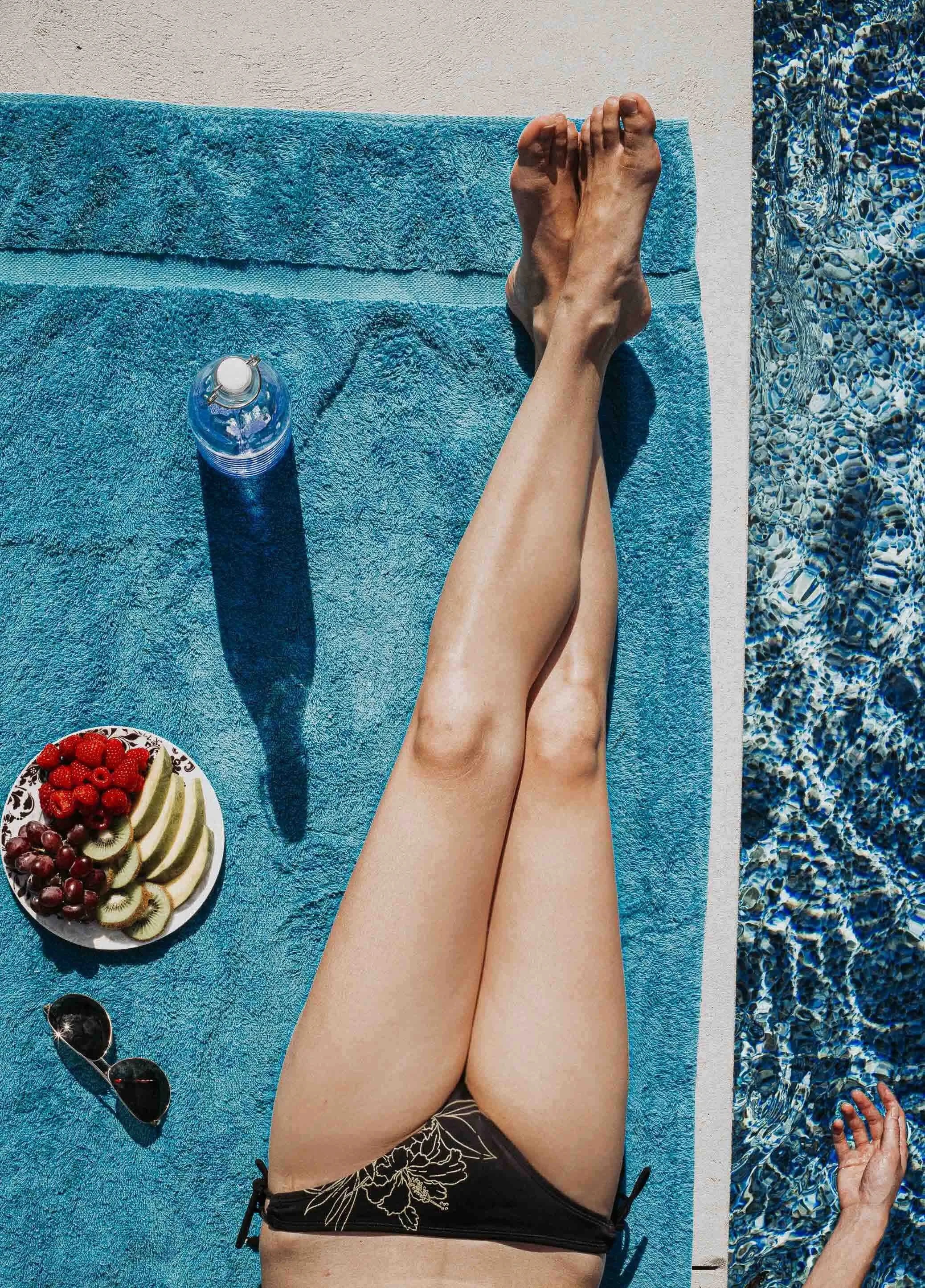Une personne allongée sur une serviette bleue au bord d'une piscine, portant un bikini noir avec des motifs floraux, avec une assiette de fruits comprenant des kiwis, des fraises, des raisins et des tranches de kiwi, une bouteille d'eau, et des lunet