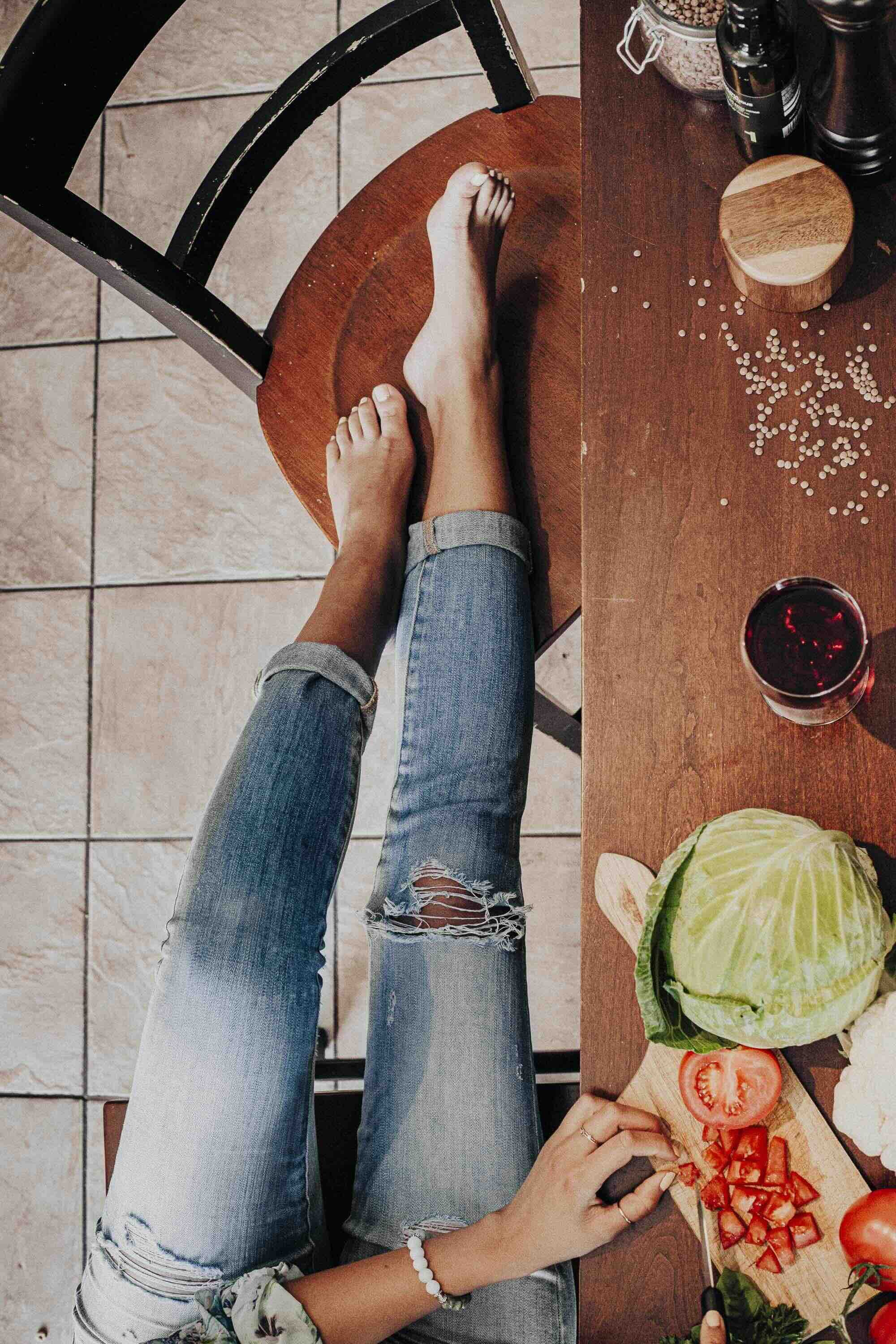 Une personne portant un jeans déchiré est assise à une table en bois, avec des légumes coupés, des bouteilles, du vin rouge et des épices sur la table.