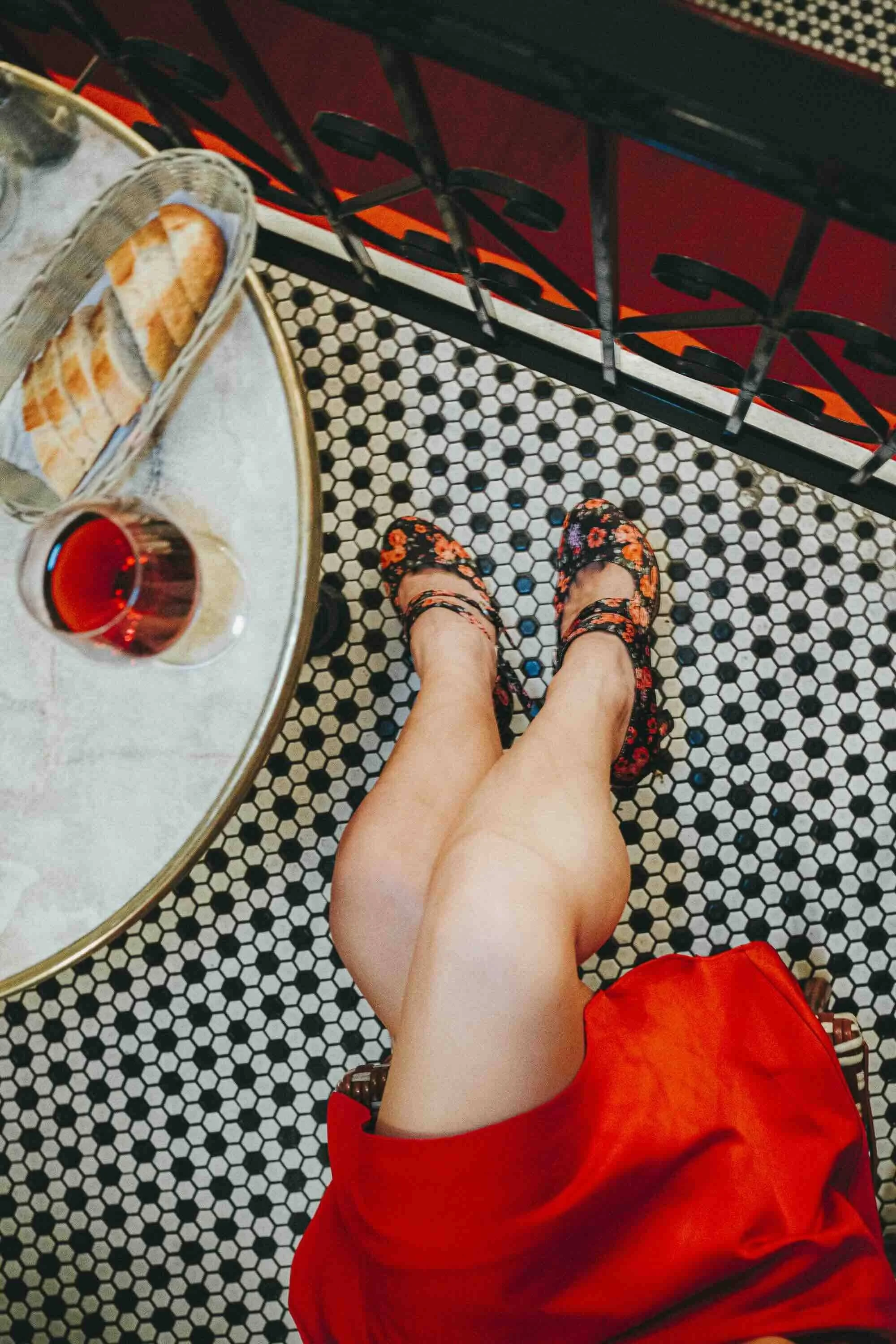 Une personne assise à une table de café, vue du dessus, portant des chaussures à talons à fleurs, une robe rouge, avec un verre de boisson rouge et une assiette de pain sur la table, un motif de carreaux noirs et blancs sur le sol.