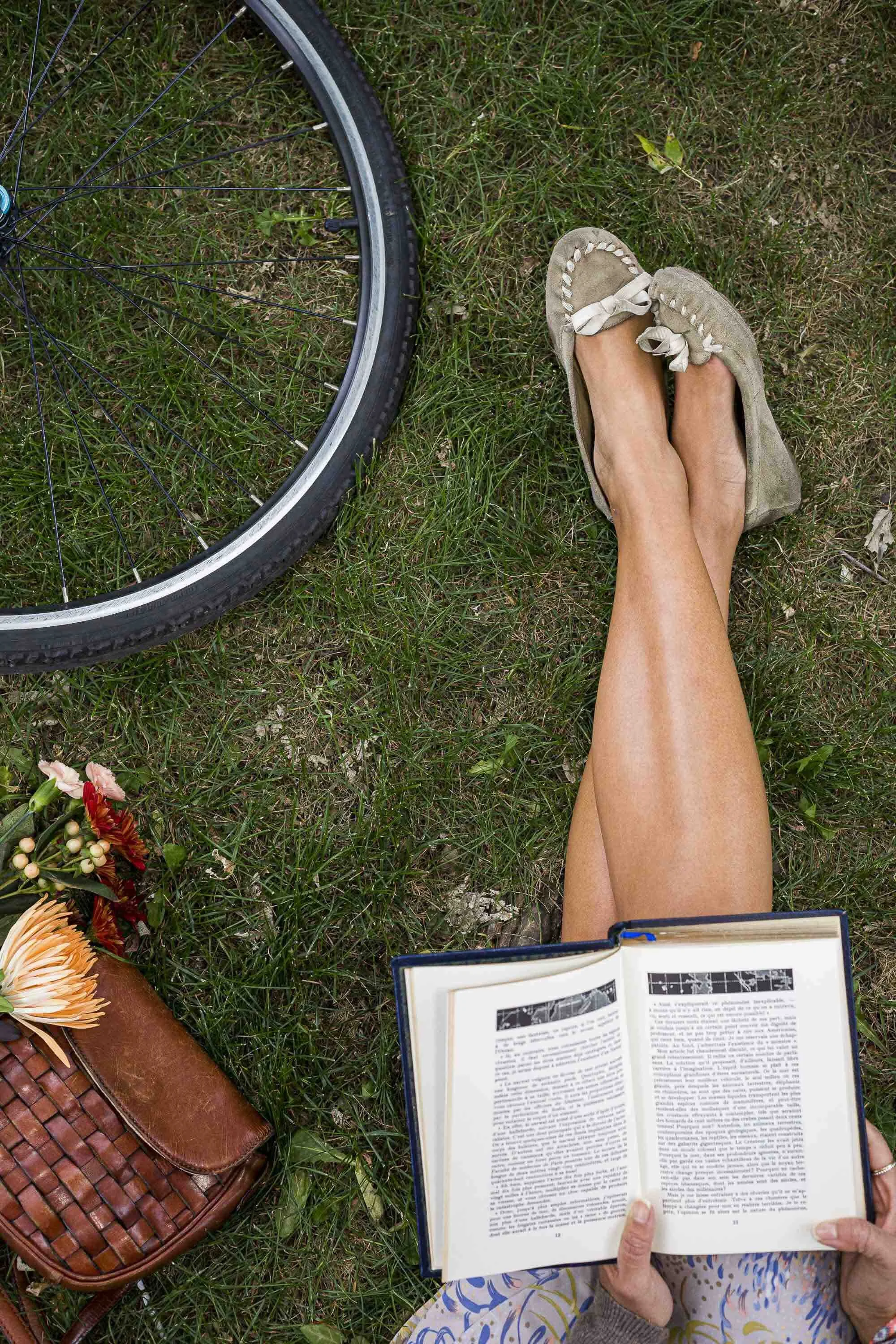 Personne assise sur l'herbe lisant un livre, avec une bicyclette à côté et un sac en cuir avec des fleurs.
