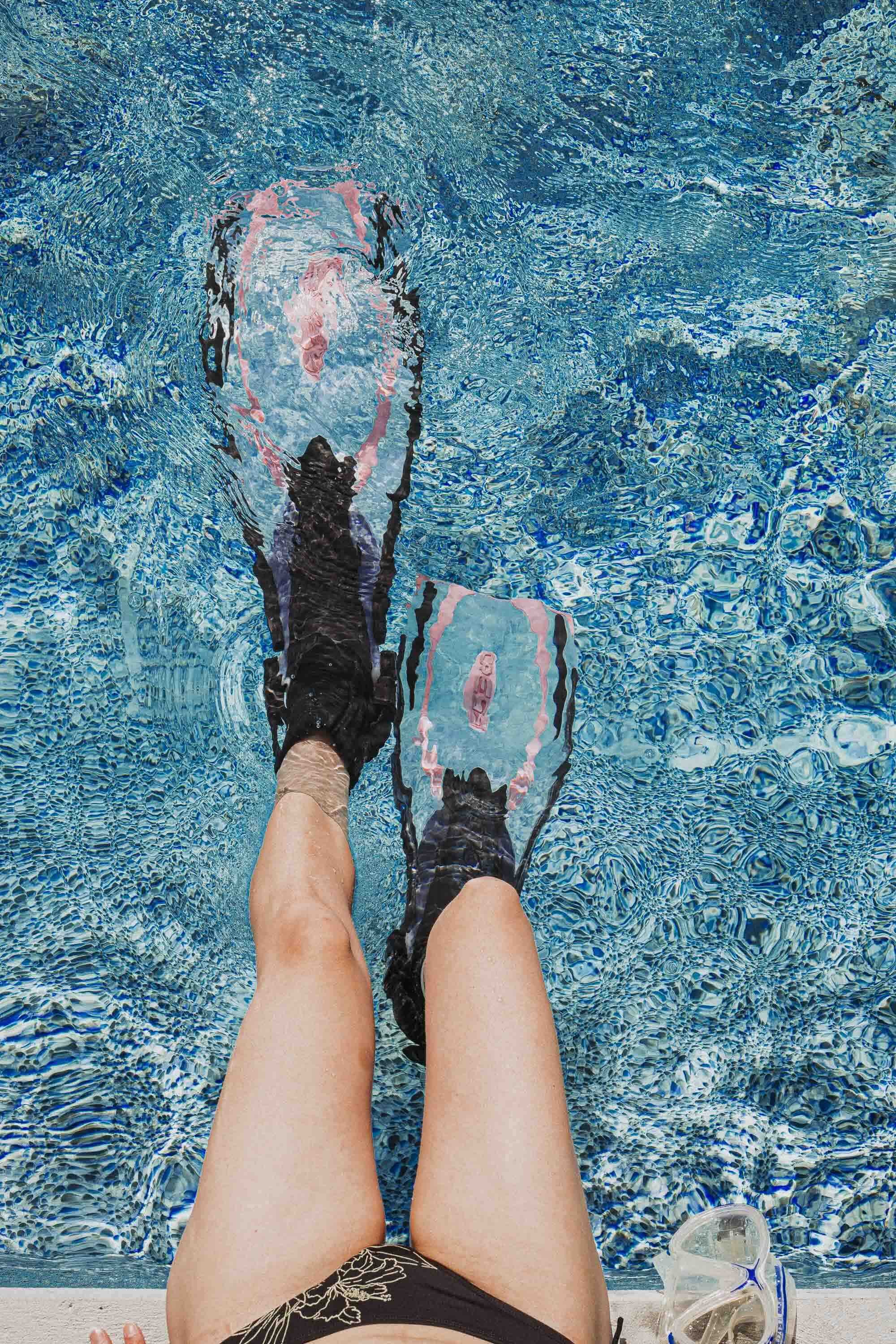 Une personne portant des chaussettes ou des chaussures d'eau dans une piscine, vue des jambes et des pieds vus de dessus, avec un fond d'eau bleue et une paire de lunettes de piscine à côté.