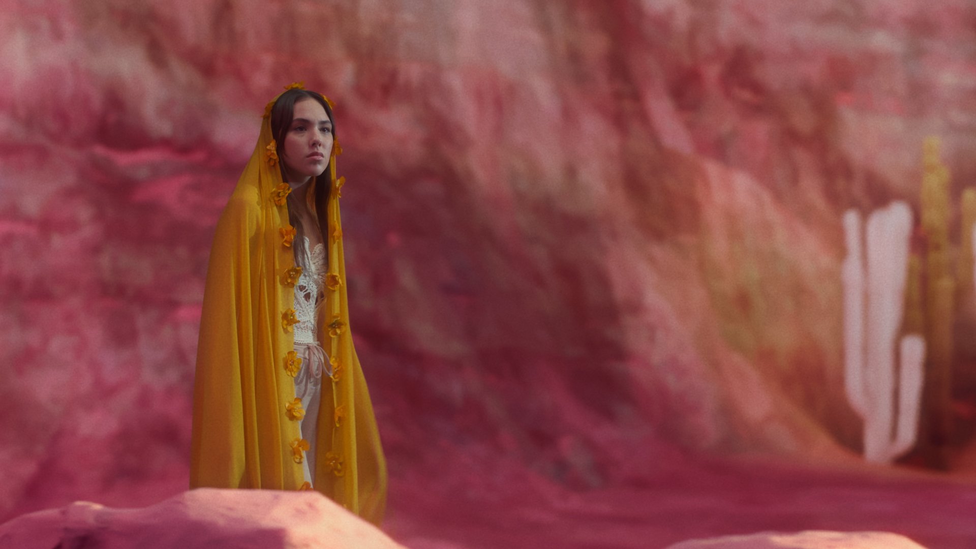 Une jeune femme portant une cape jaune avec des fleurs, debout dans un décor de couleur rose et rouge, semblable à un paysage fantastique ou une scène de film.
