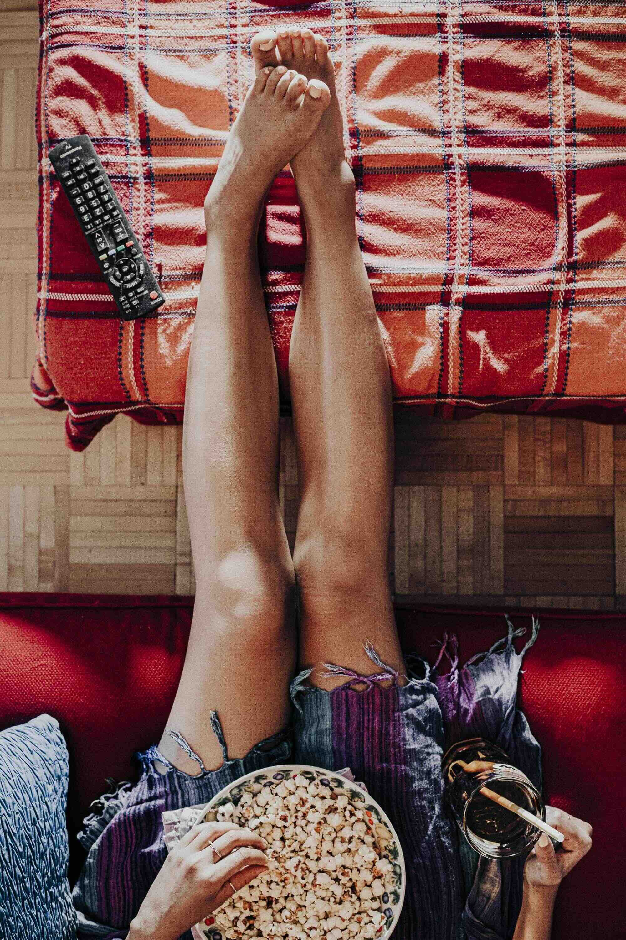 Une personne assise sur un canapé rouge, tenant un bol de popcorn dans une main et une boisson dans l'autre, avec des jambes allongées sur une table recouverte d'une nappe à carreaux.