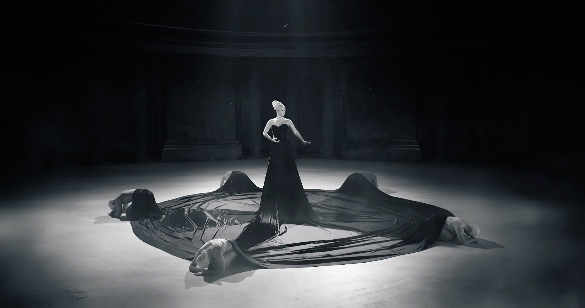 Une scène en noir et blanc avec une femme debout au centre, portant une robe longue noire, entourée de quatre personnes allongées, toutes sur une scène de théâtre ou un studio.