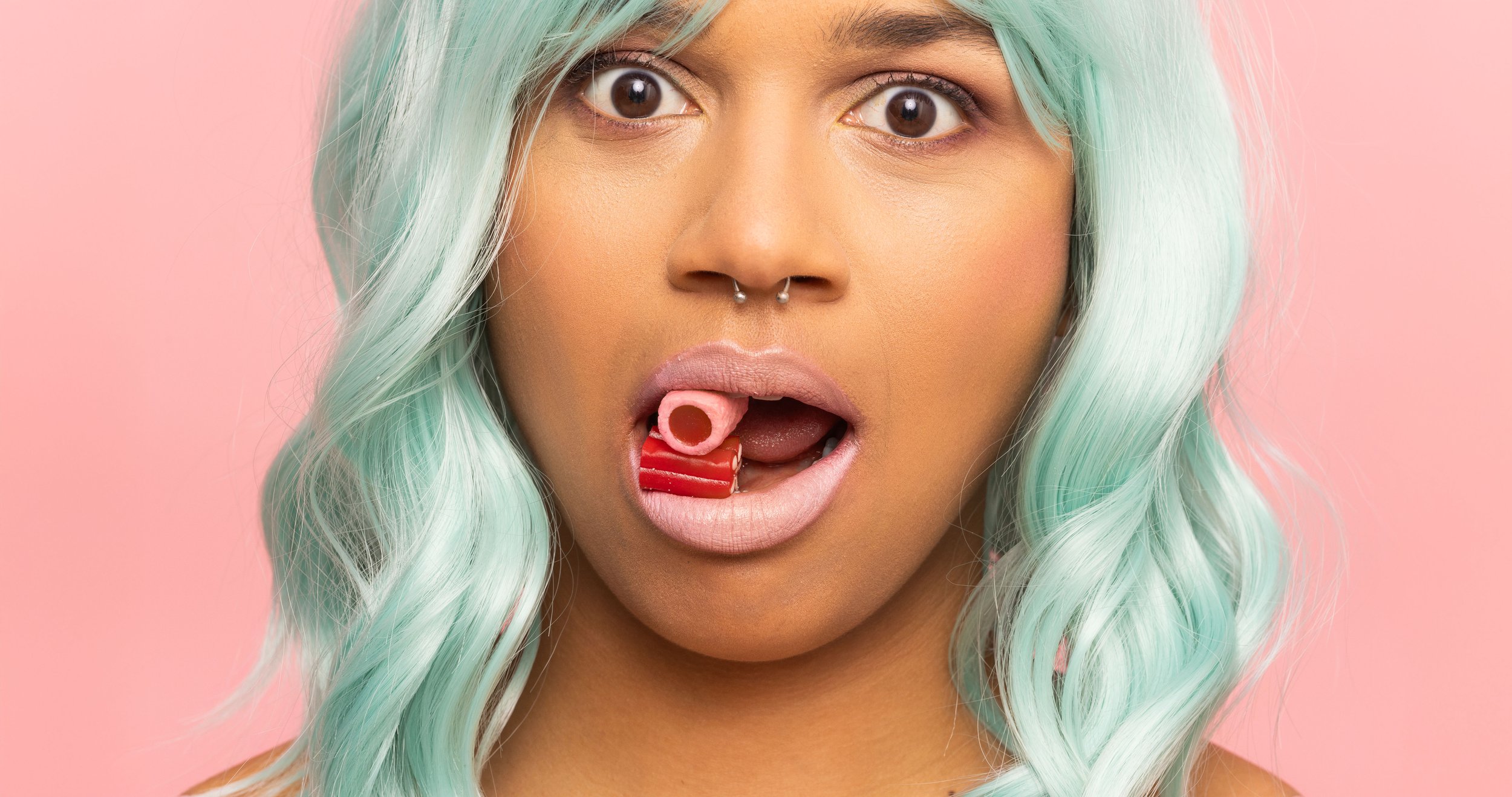 Une femme aux cheveux longs, de couleur turquoise, avec un piercing au nez, ouvre la bouche et tient un rouleau de bonbon rouge dans sa bouche, contre un fond rose.