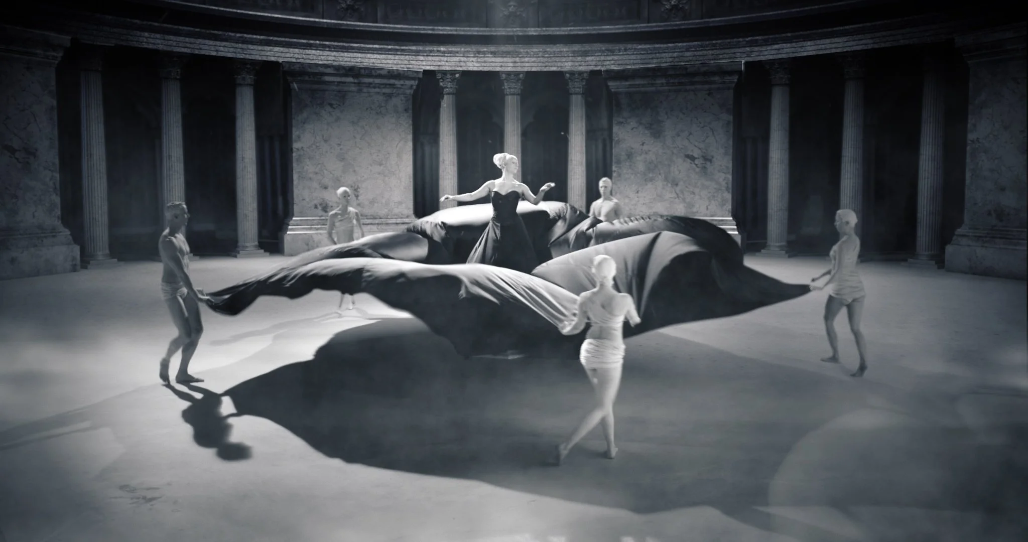 Des femmes en robe dansent avec un grand tissu noir dans une salle avec des colonnes en marbre.