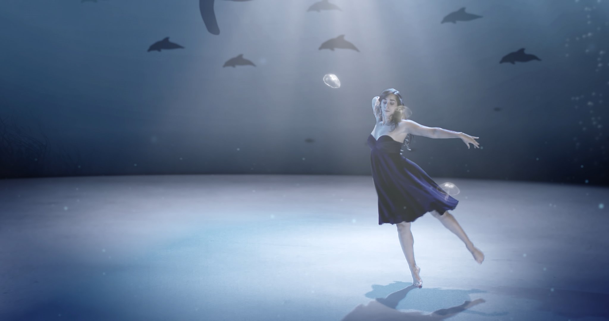 Une ballerine en danse sur scène sous un éclairage, avec des poissons nageant en arrière-plan dans une atmosphère aquatique.