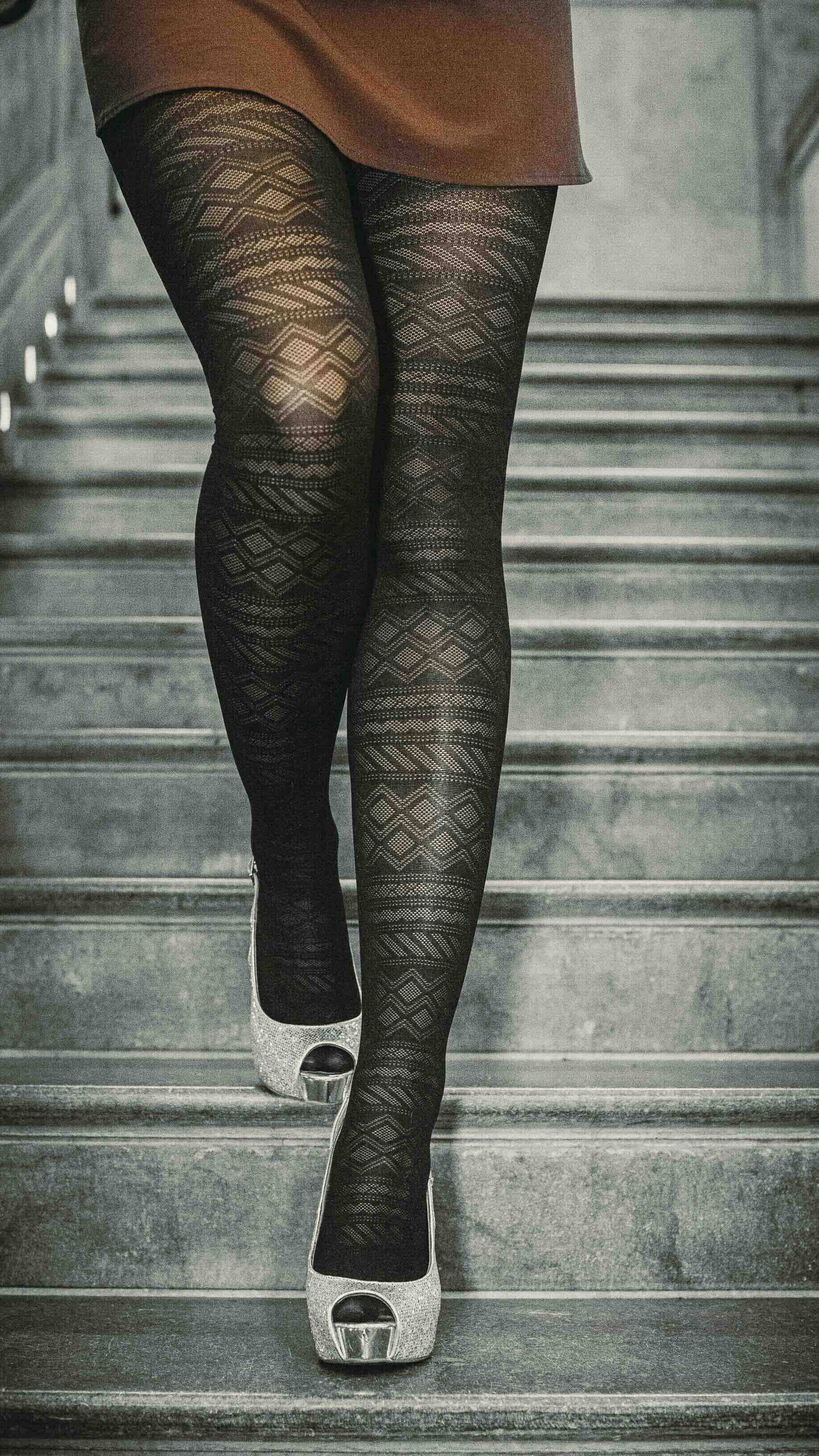 Une personne portant des collants noirs avec un motif géométrique, marchant dans une montée d'escalier en béton avec des chaussures à talons à paillettes argentées.