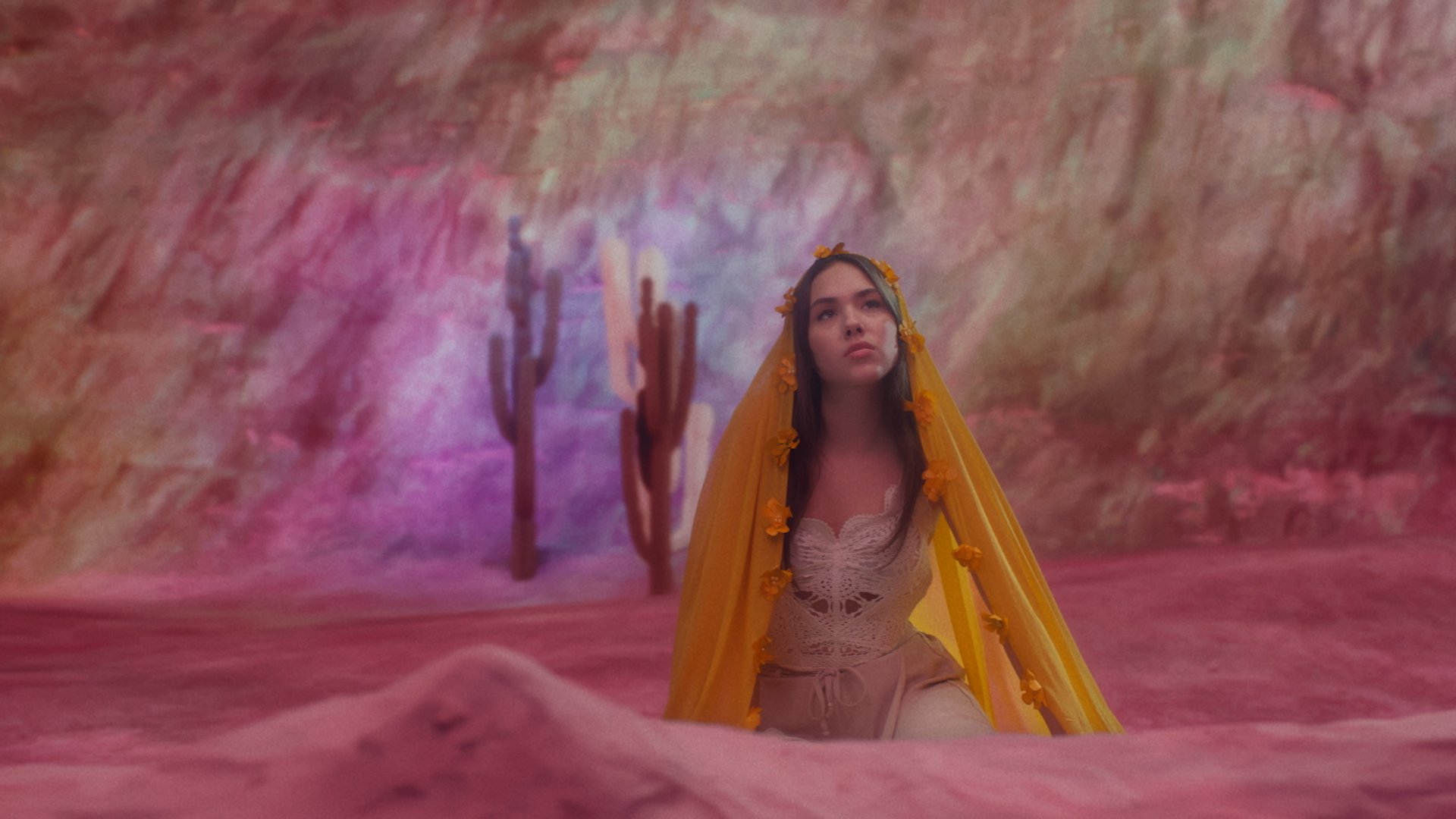 Une jeune femme vêtue d'une robe blanche et d'un voile coloré en jaune, assise dans un paysage coloré avec des cactus en arrière-plan.