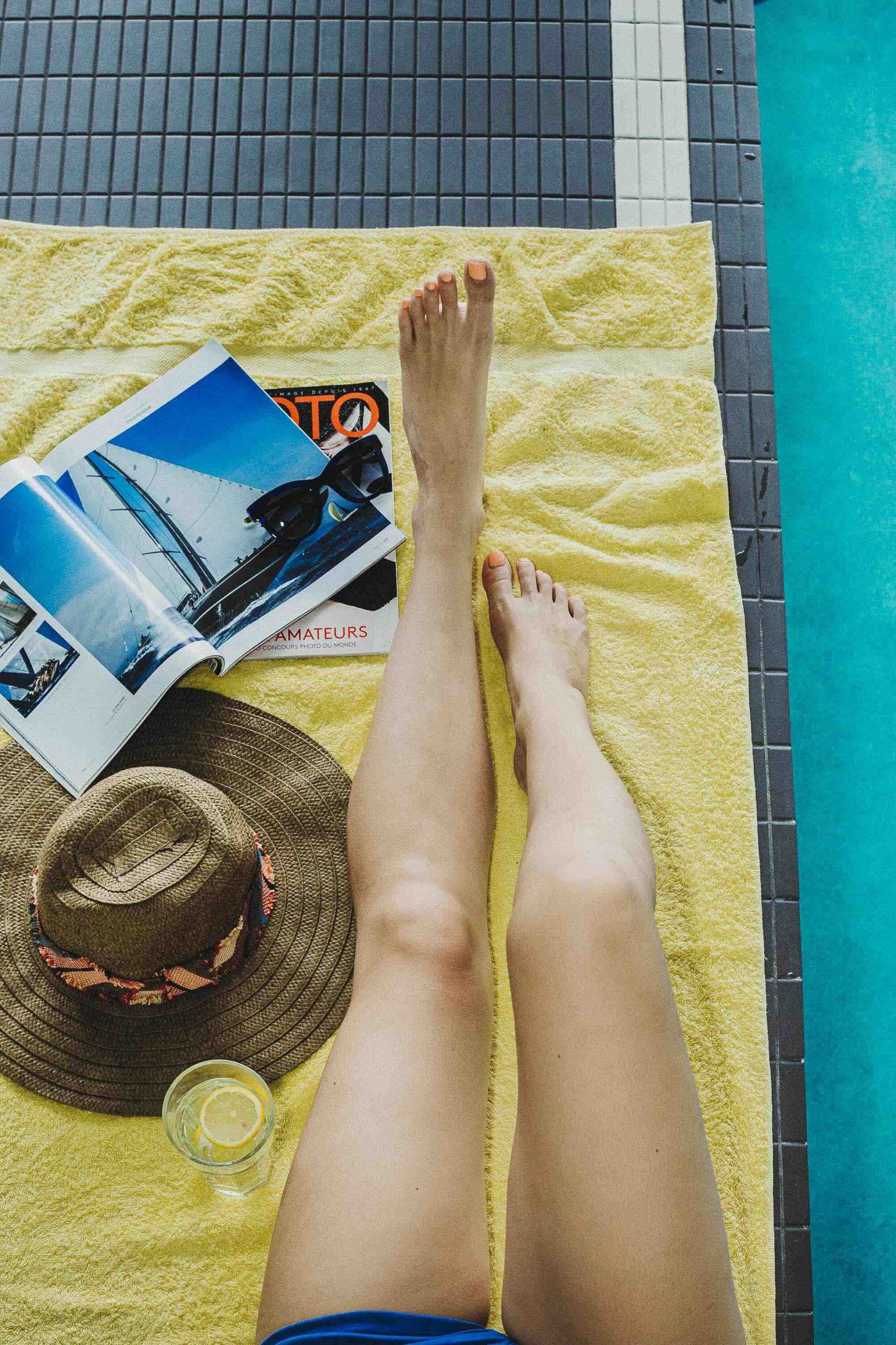 Une personne est allongée sur une serviette jaune près d'une piscine, avec un magazine, un chapeau en paille, une boisson avec une tranche de citron, et un livre ouvert.